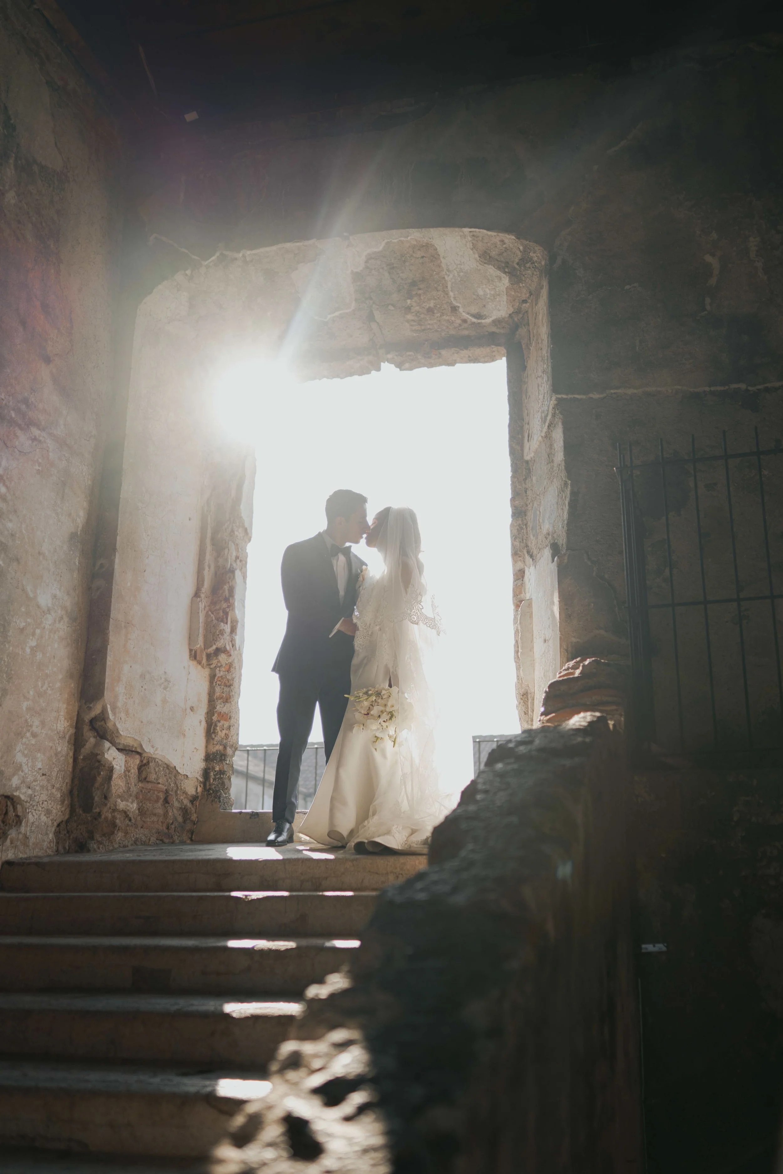 Pareja de novios en una boda con luz brillante y fondo desaturado, posando en una puerta de piedra antigua.