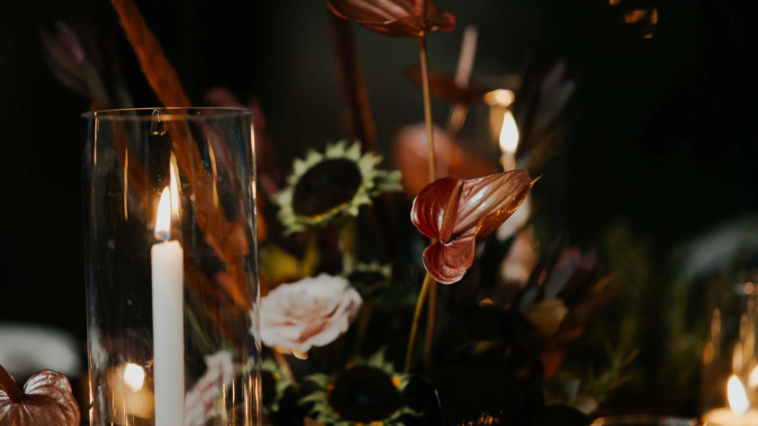 Velas encendidas entre flores de diferentes colores y tipos en un fondo oscuro.