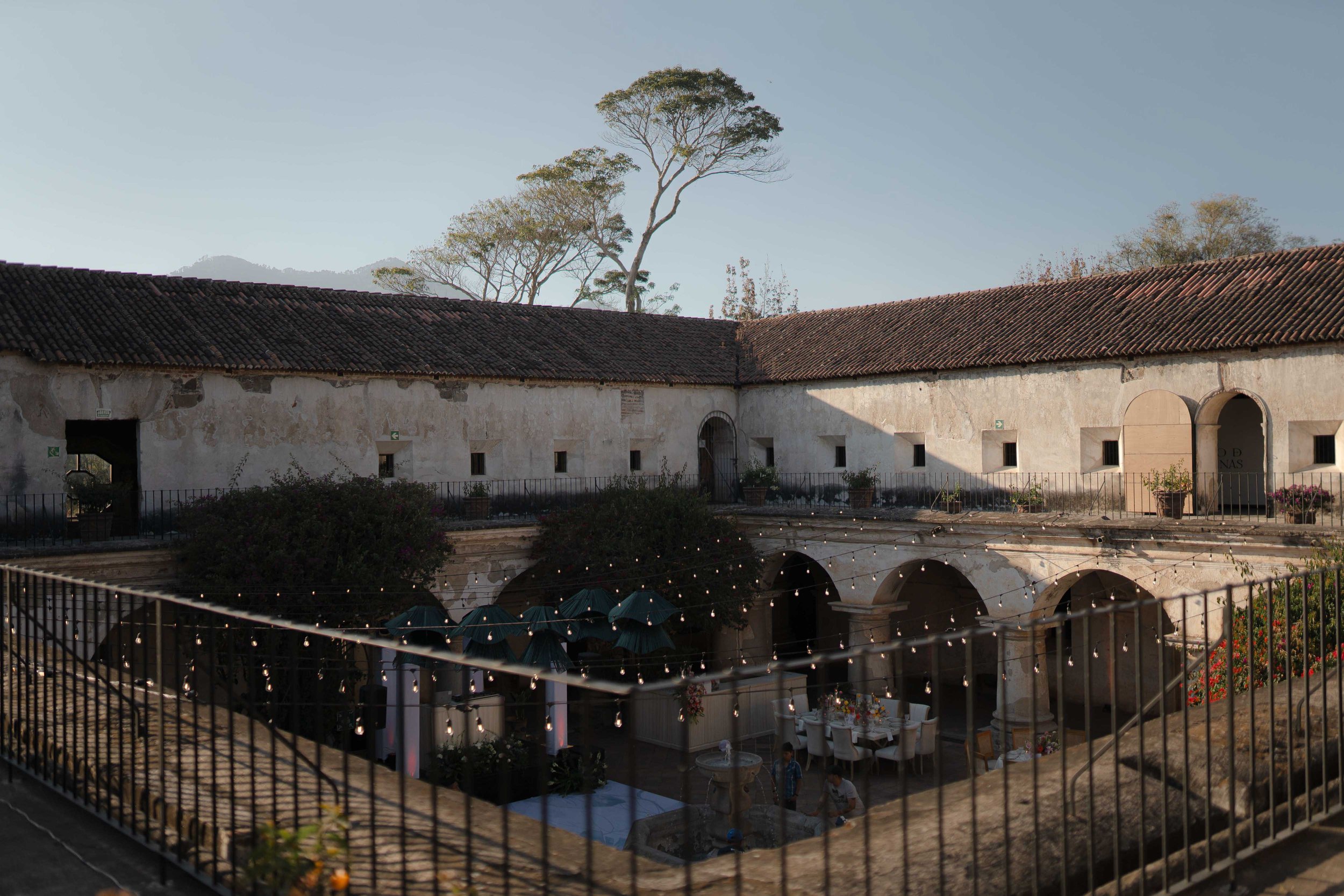 Vista de un patio interior de un edificio antiguo con paredes de piedra y un techo de tejas, decorado con luces colgantes, mesas y sombrillas, y árboles en el fondo.