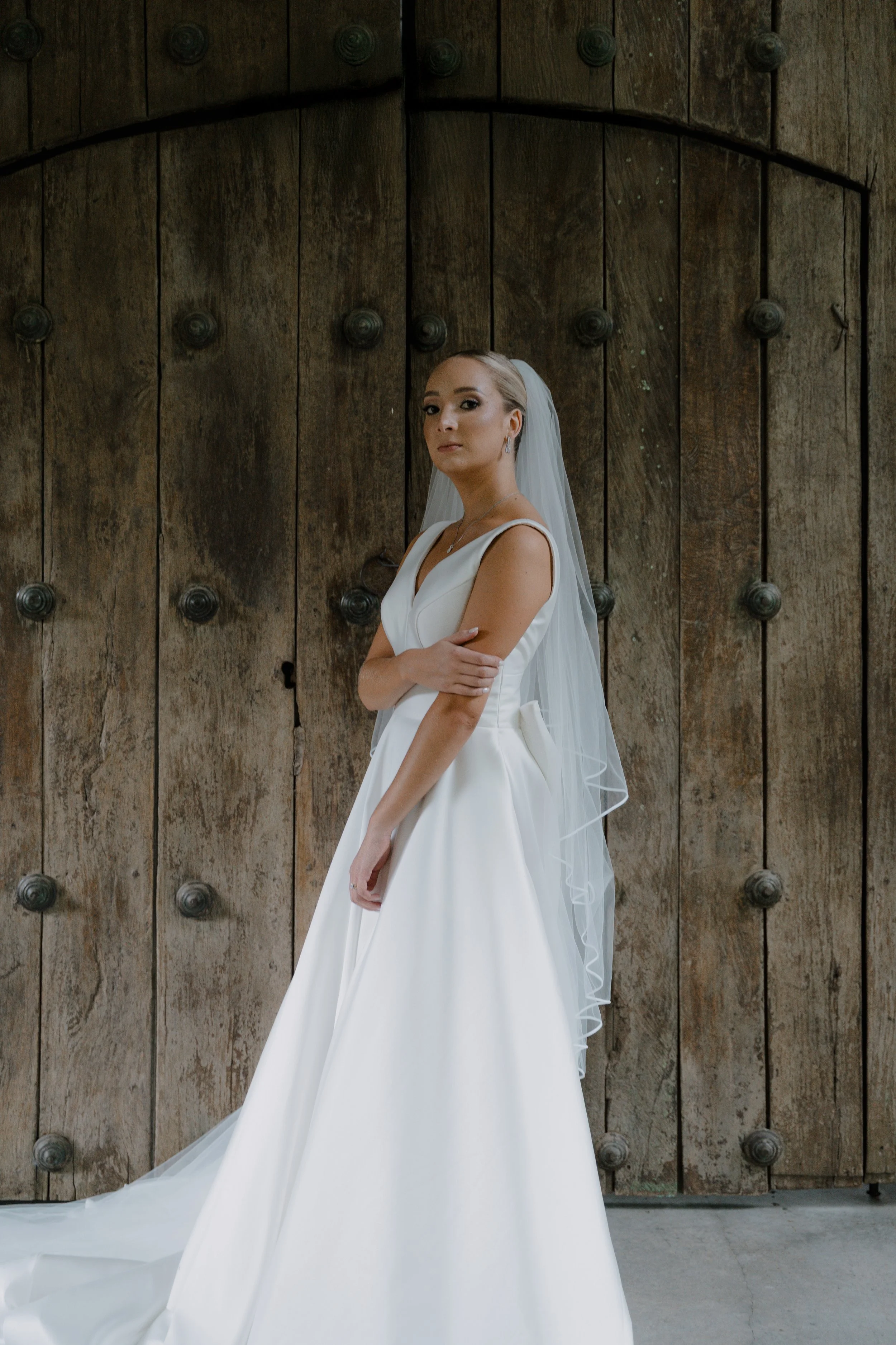 Mujer con vestido de novia blanco y velo en frente de una gran puerta de madera antigua.
