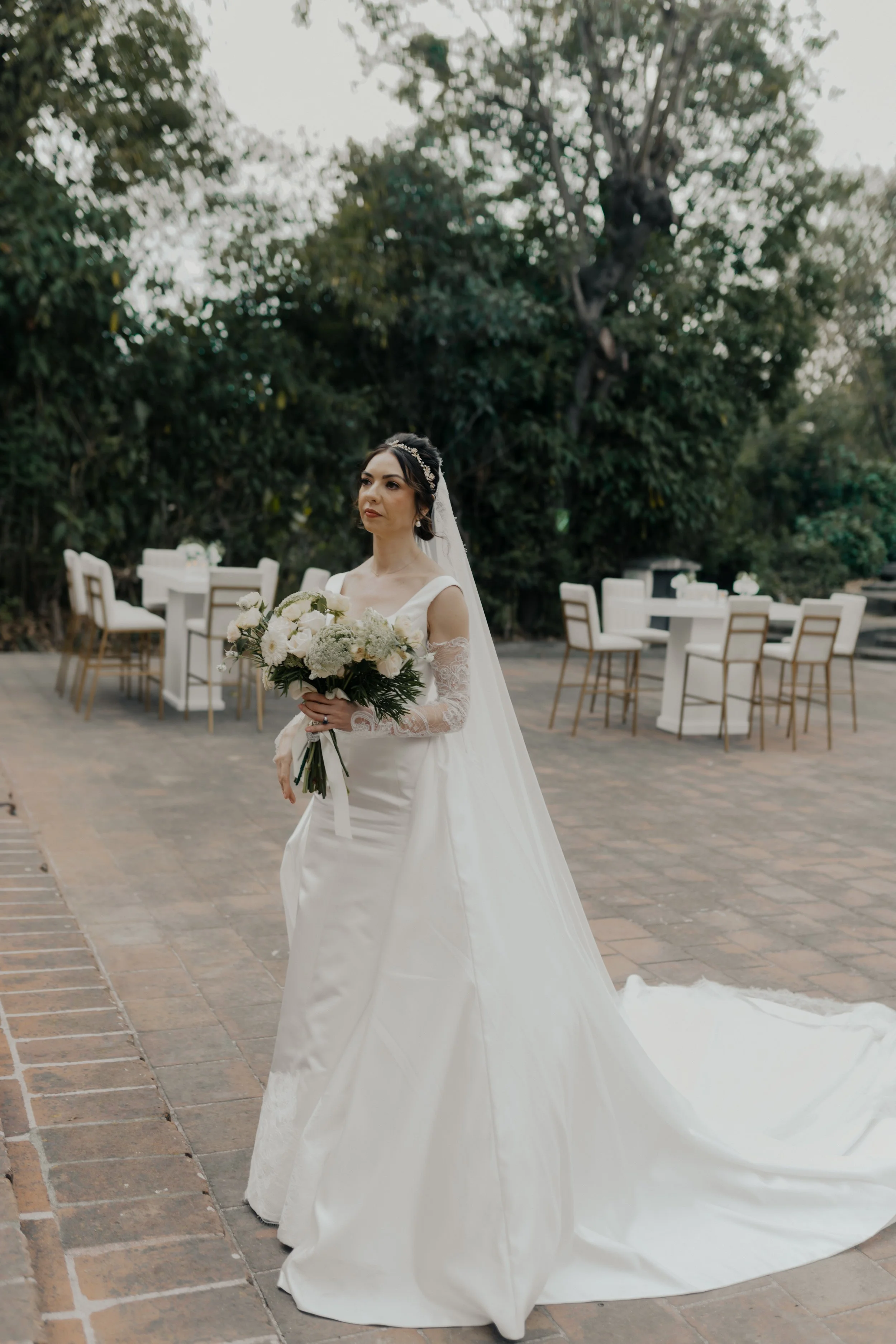 Mujer vestida de novia con ramo en una ceremonia al aire libre, rodeada de árboles y mesas con sillas blancas.