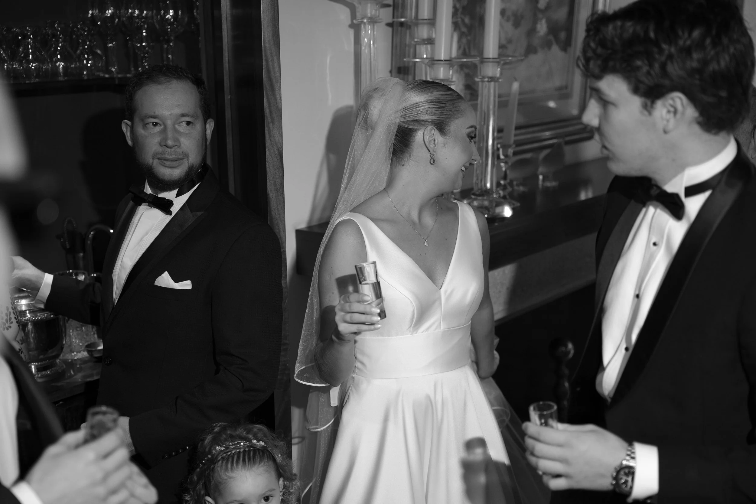 Pareja de novios en una fiesta, la novia viste vestido blanco y el novio traje negro; una mujer en vestido blanco de pie, sosteniendo una copa, sonriendo, y un hombre con barba en un traje de etiqueta en una reunión social en interiores.
