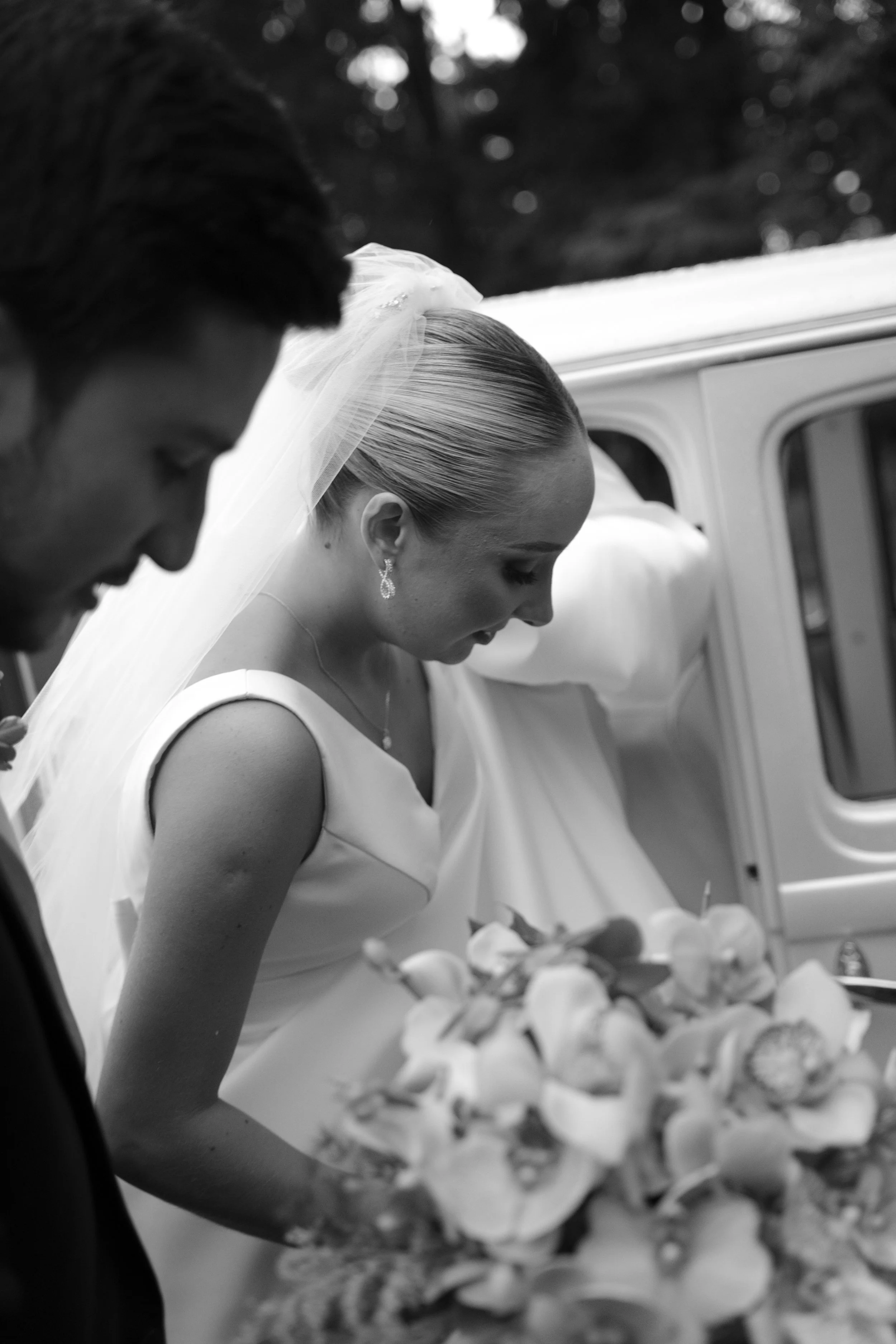 Una mujer en vestido de novia y un hombre en ropa formal, ambos inclinados hacia abajo, en un momento de la ceremonia de boda, con un coche viejo de fondo y un ramo de flores en primer plano.