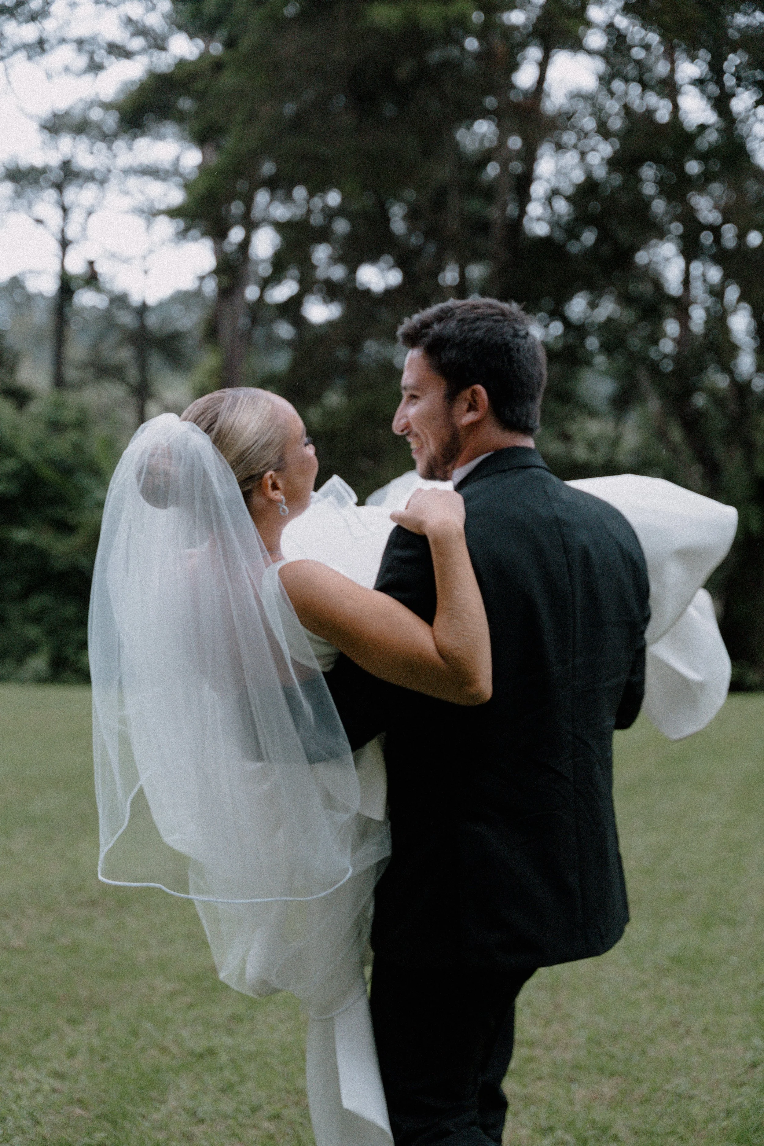 Una pareja de novios bailando en un parque, ella con un velo y él con un traje negro, celebrando su boda al aire libre con árboles en el fondo.