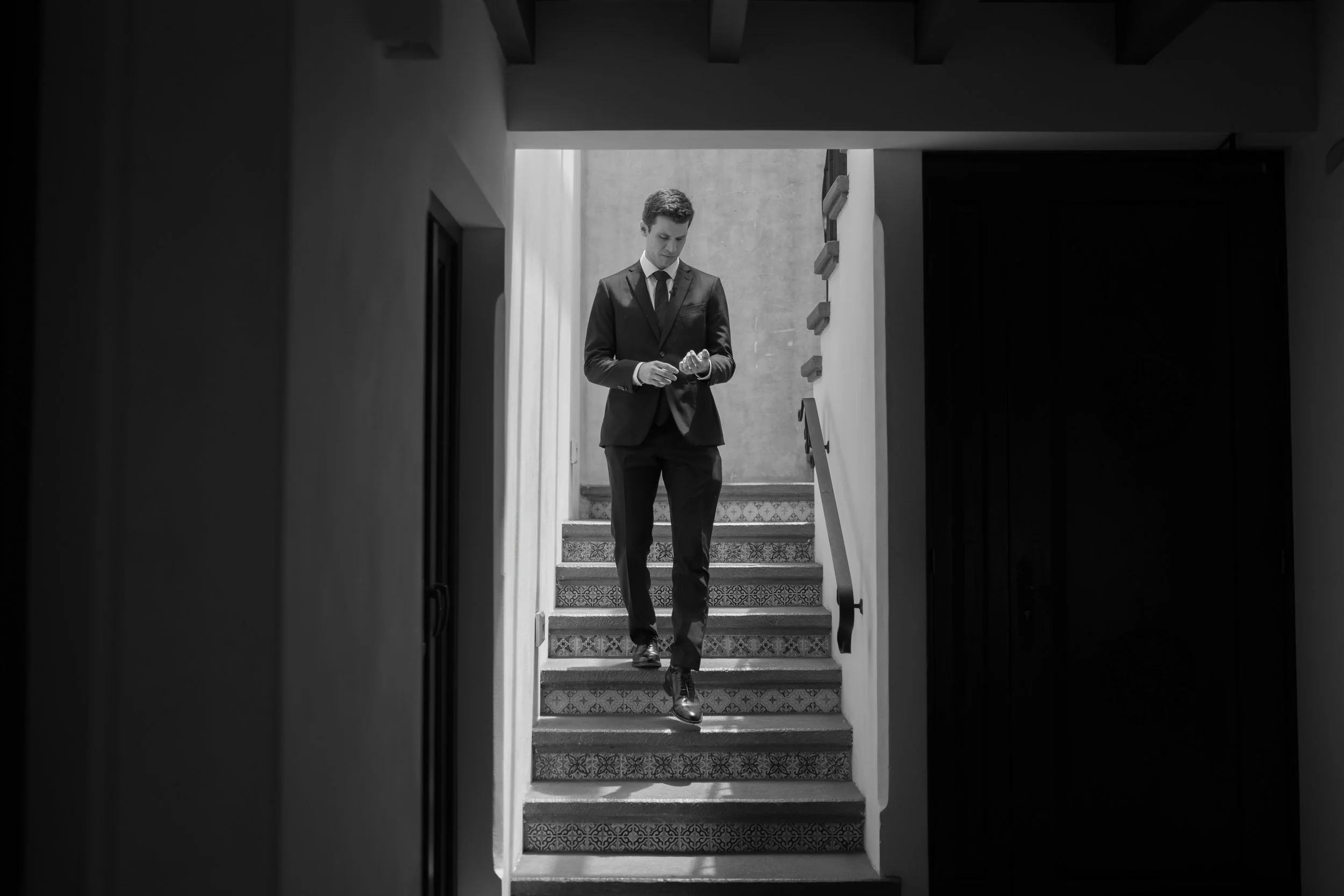 Hombre en traje oscuro bajando escaleras de una casa, mirando su reloj, en una fotografía en blanco y negro.