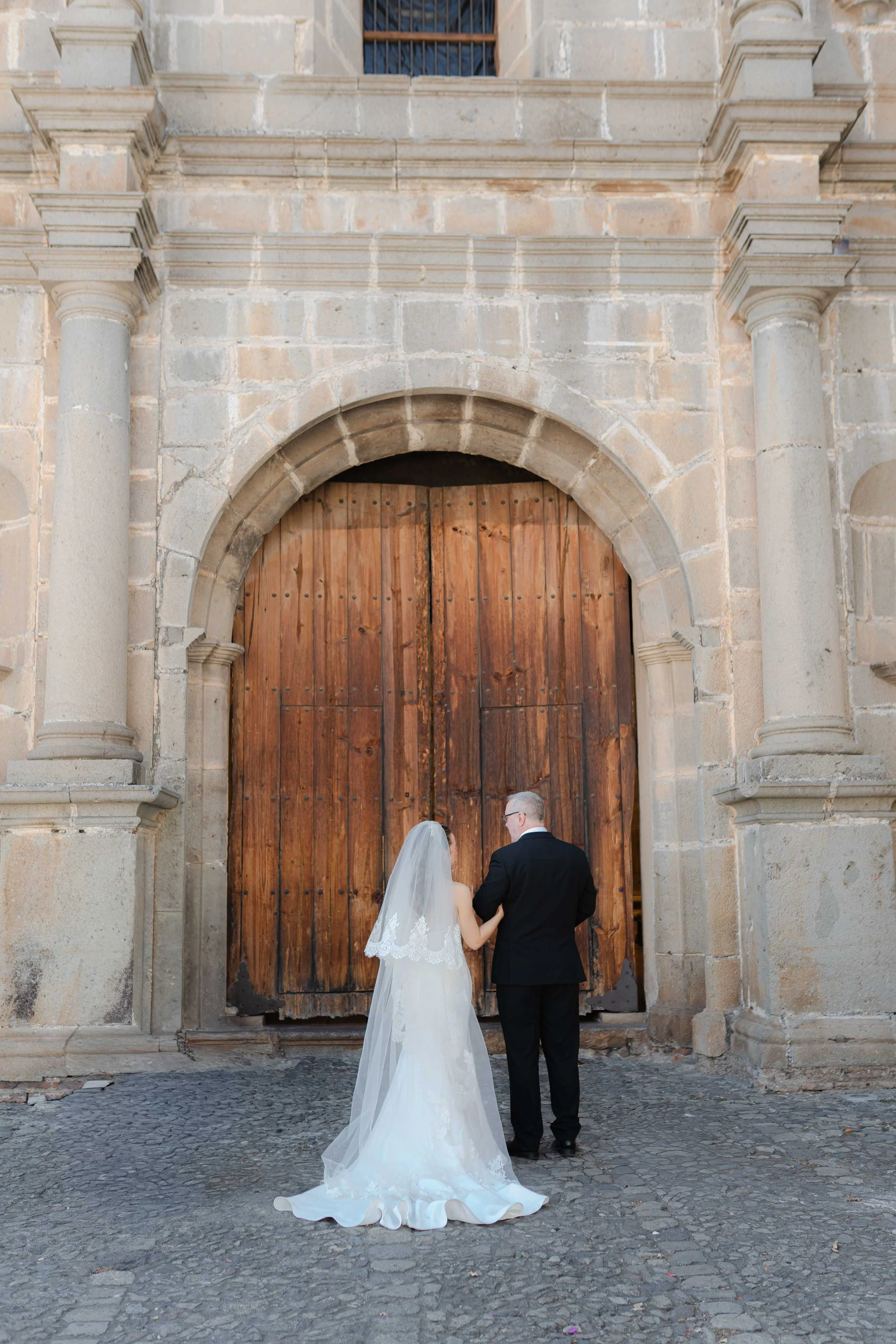 Pareja de novios de espaldas, la novia con vestido blanco y velo, el novio con traje negro, frente a una gran puerta de madera en una iglesia antigua de piedra.