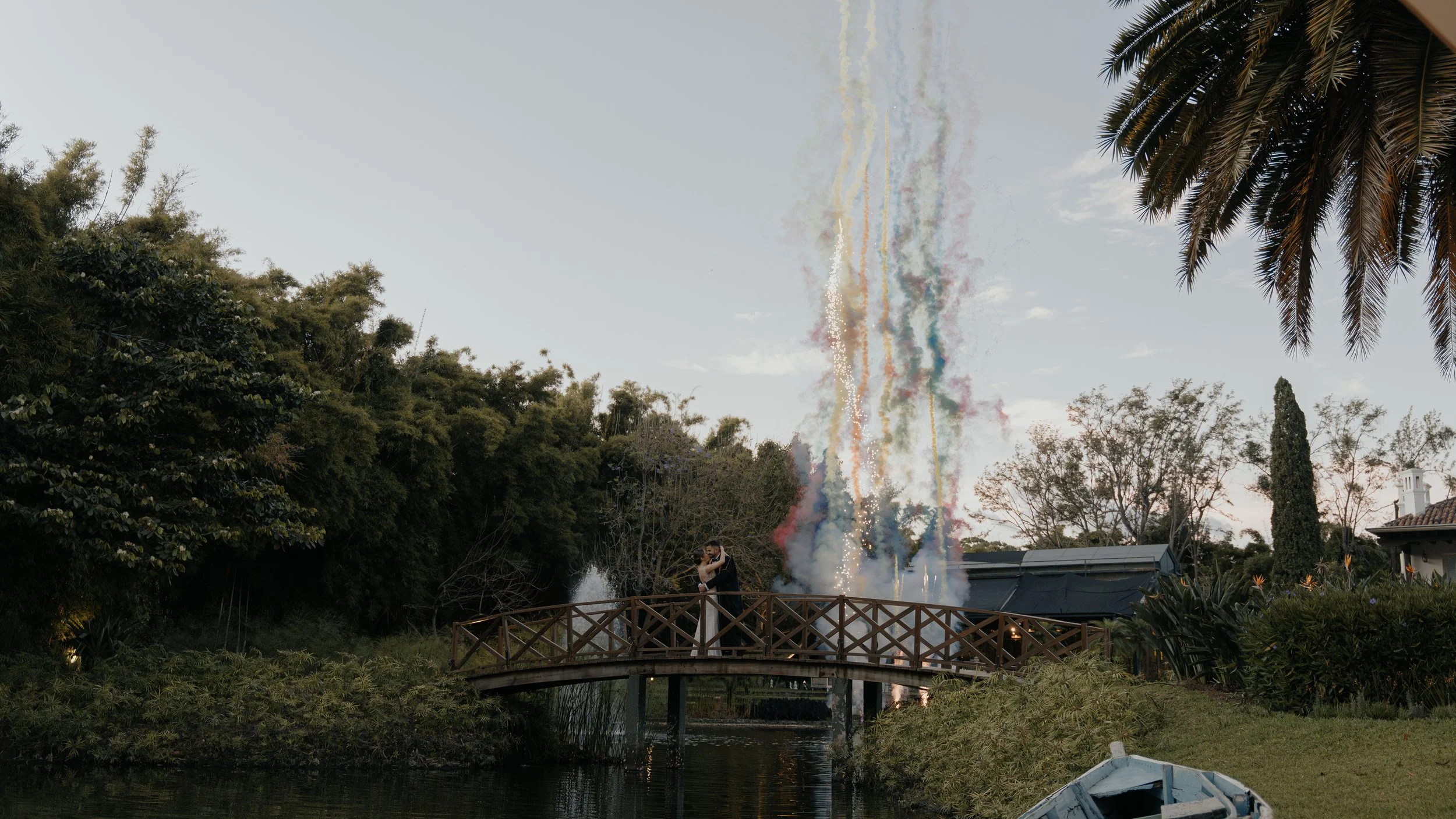 Pareja besándose en un puente de madera sobre un cuerpo de agua, con fuegos artificiales en el fondo y árboles a su alrededor.