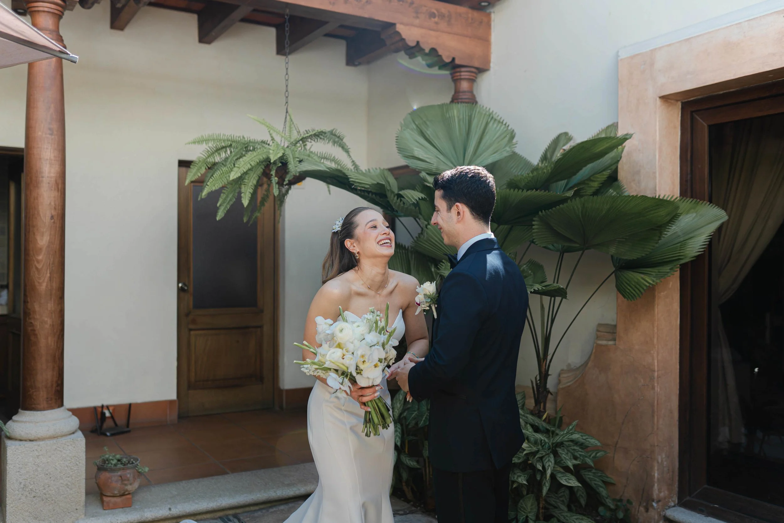 Pareja de recién casados riendo y mirándose en un jardín decorado con plantas grandes y una puerta de madera al fondo.