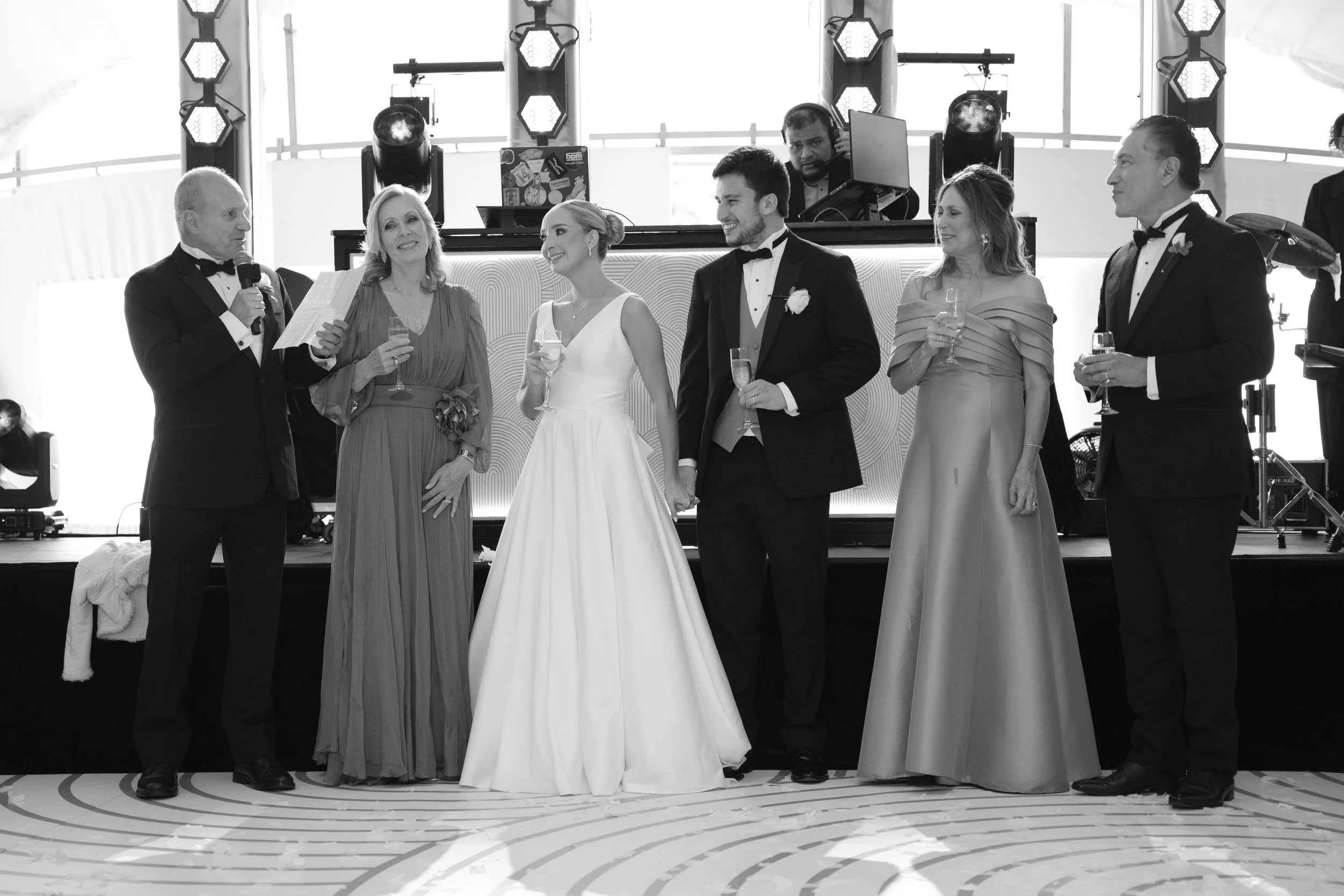Grupo de personas en un evento formal, algunos con ropa de fiesta y sosteniendo copas, en un escenario con luces y un DJ detrás, en un ambiente de celebración.