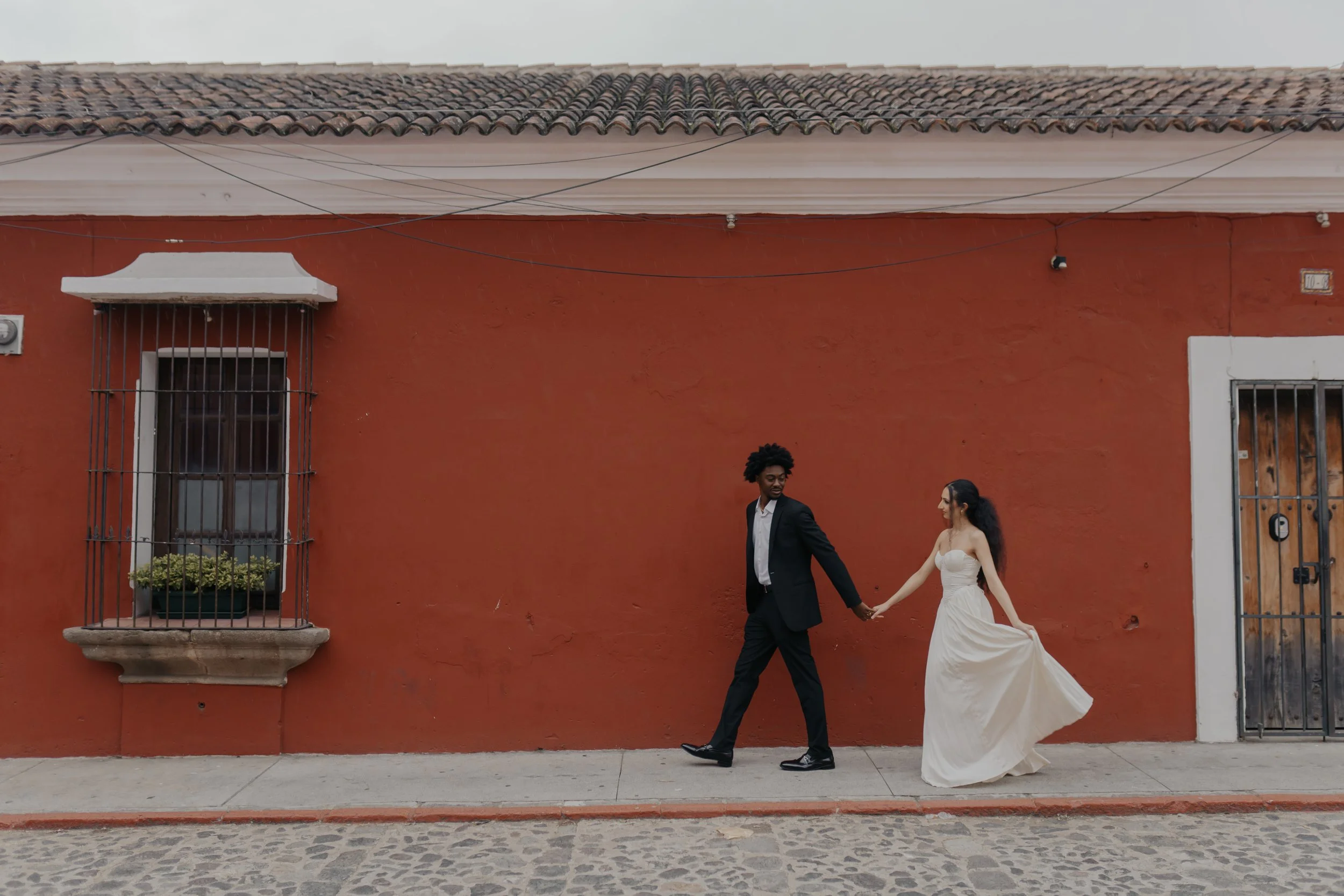 Una pareja vestida formalmente, caminando de la mano en una calle de adoquines frente a una pared anaranjada, la mujer lleva un vestido largo blanco y el hombre lleva un traje negro.