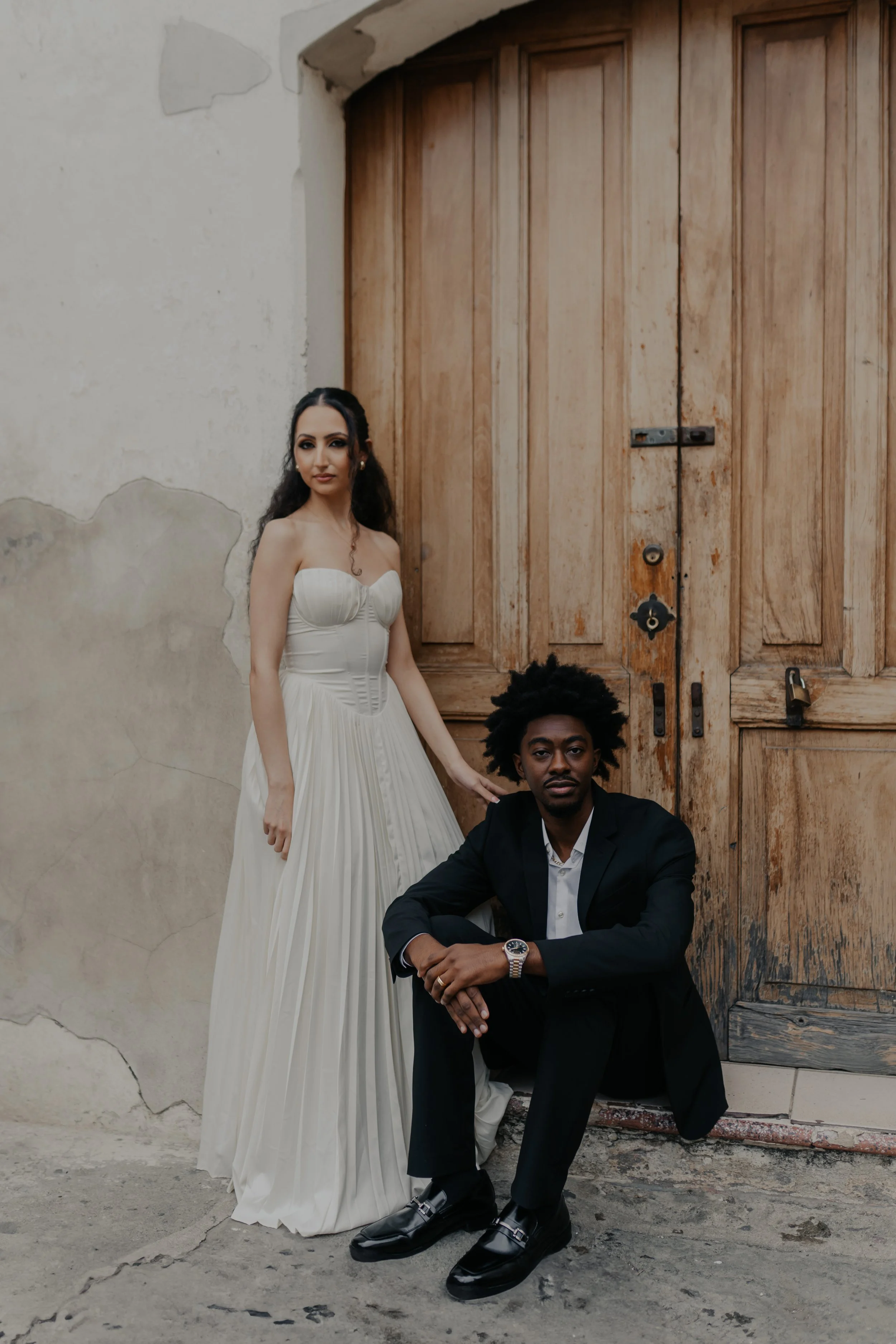 Una pareja elegante posa frente a una puerta de madera, la mujer lleva vestido blanco y el hombre traje oscuro.