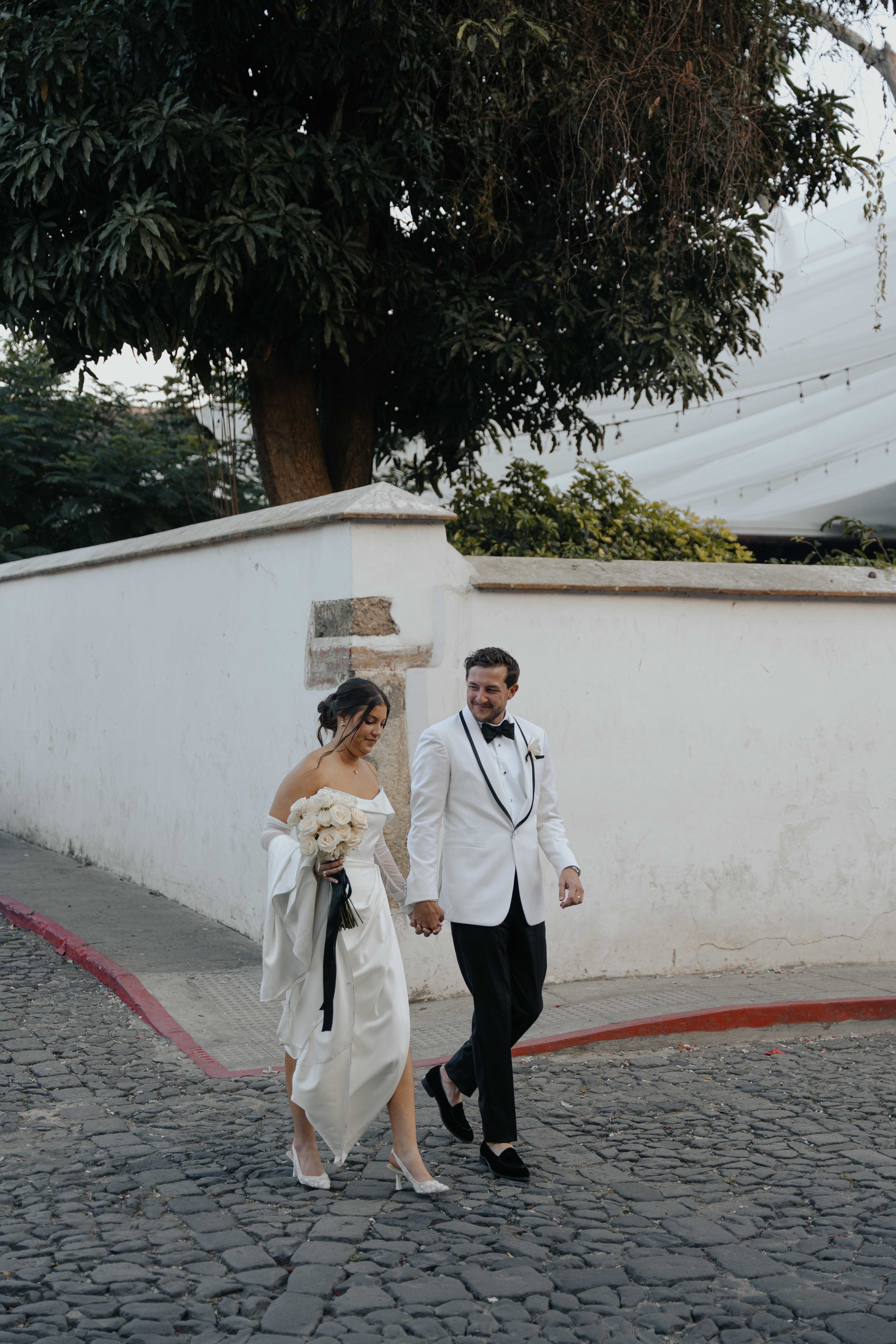 Pareja de boda caminando por la calle de adoquines, ella con vestido blanco y ramo de rosas, él con saco blanco y pajarita, sosteniéndose de las manos.