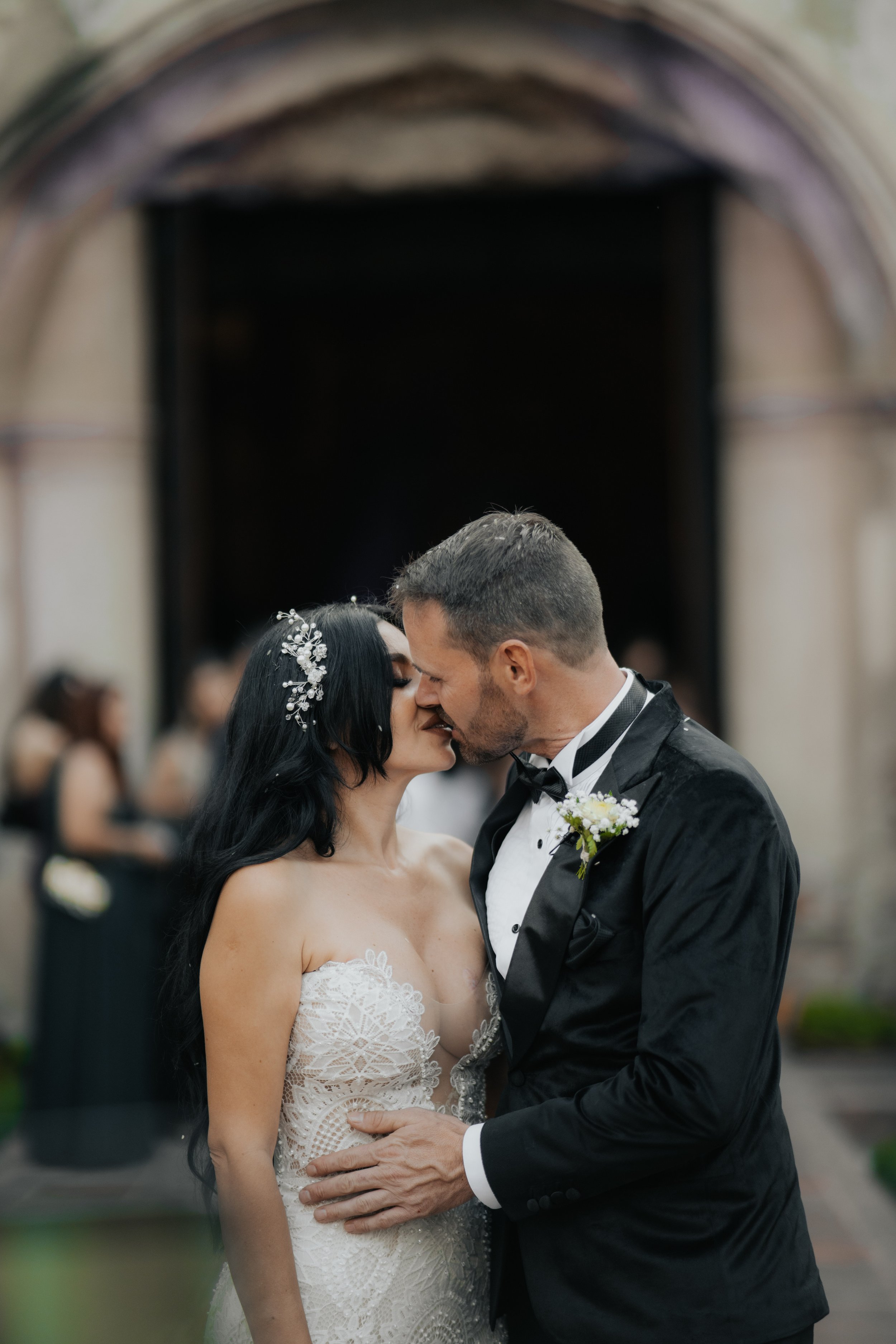 Pareja de recién casados besándose en la ceremonia en un lugar con arquitectura antigua y un arco de piedra, con invitados al fondo.