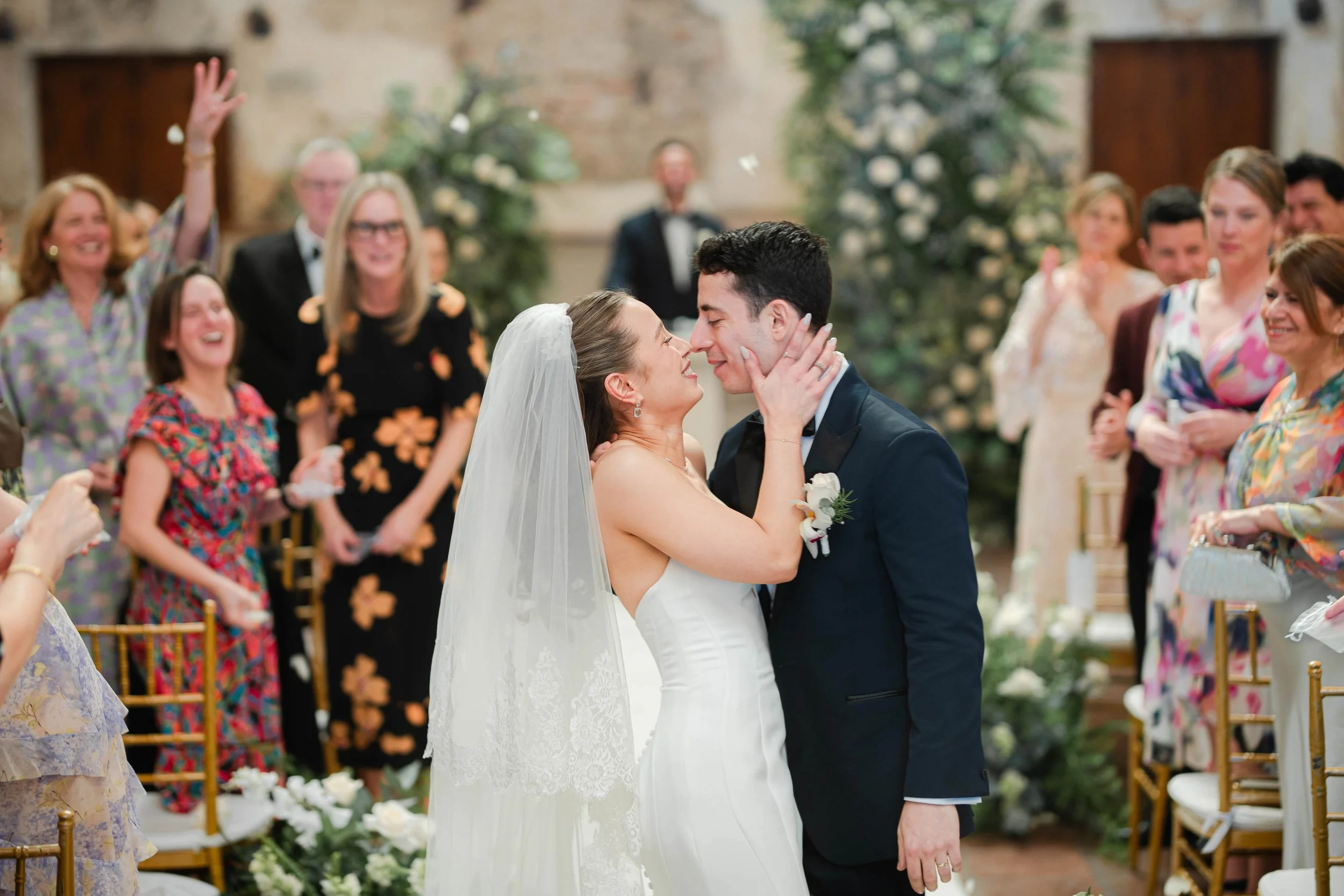 Pareja de novios en su boda, tocándose la cara y sonriendo, rodeados de invitados en un salón decorado con flores.