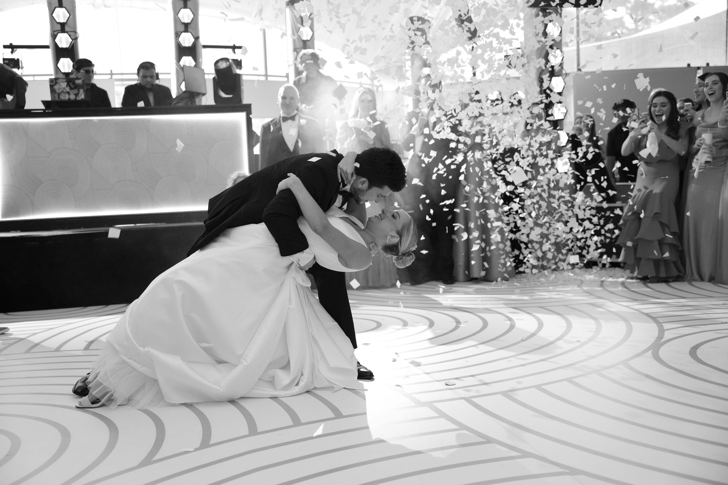 Una pareja baila en su boda, la pareja está en un abrazo con sonrisa, rodeados de invitados y confeti en el aire.