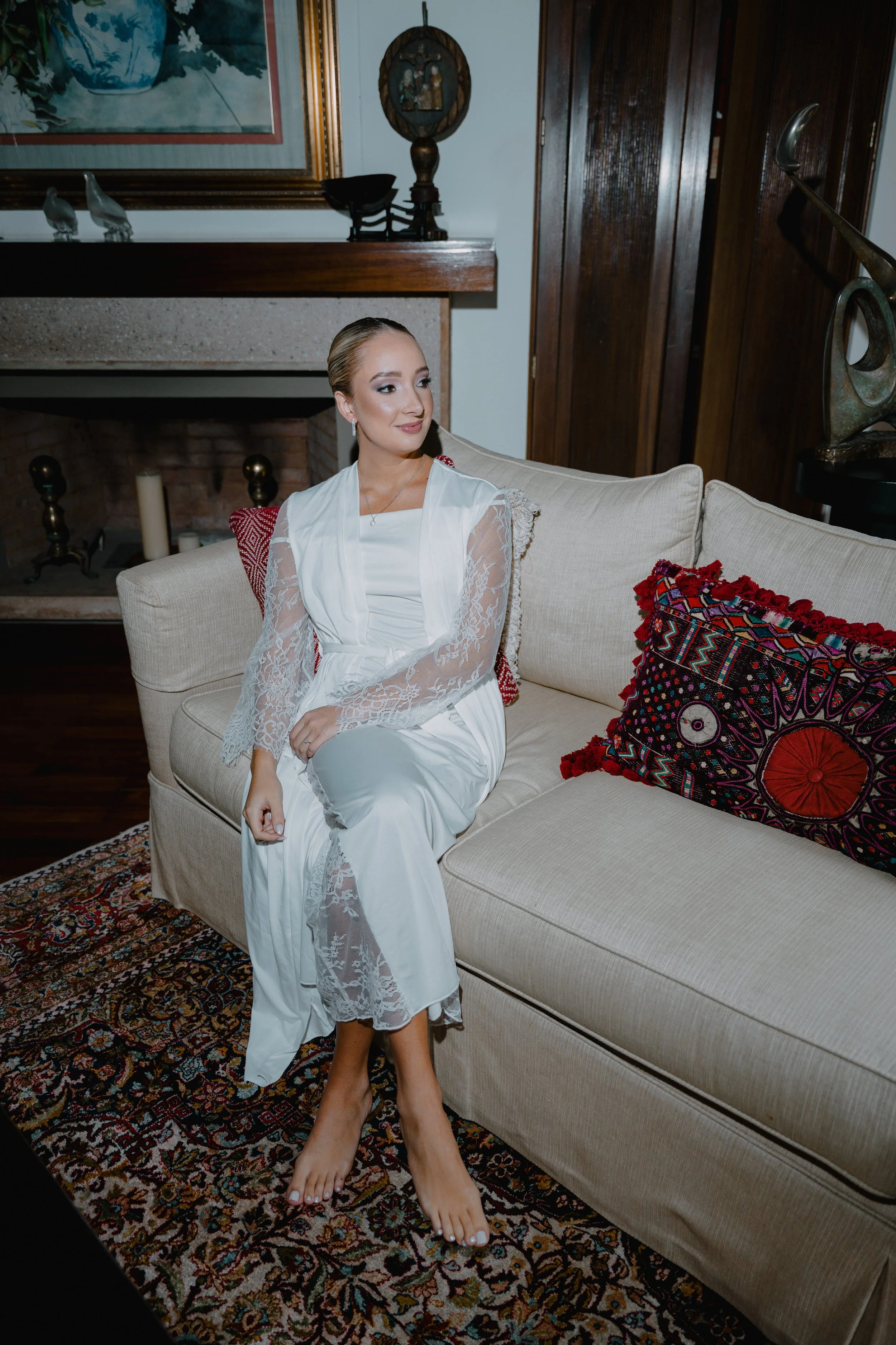Joven mujer con vestido blanco de encaje, sentada en un sofá beige, en una sala decorada con un tapiz con bordado de colores y cojines coloridos.