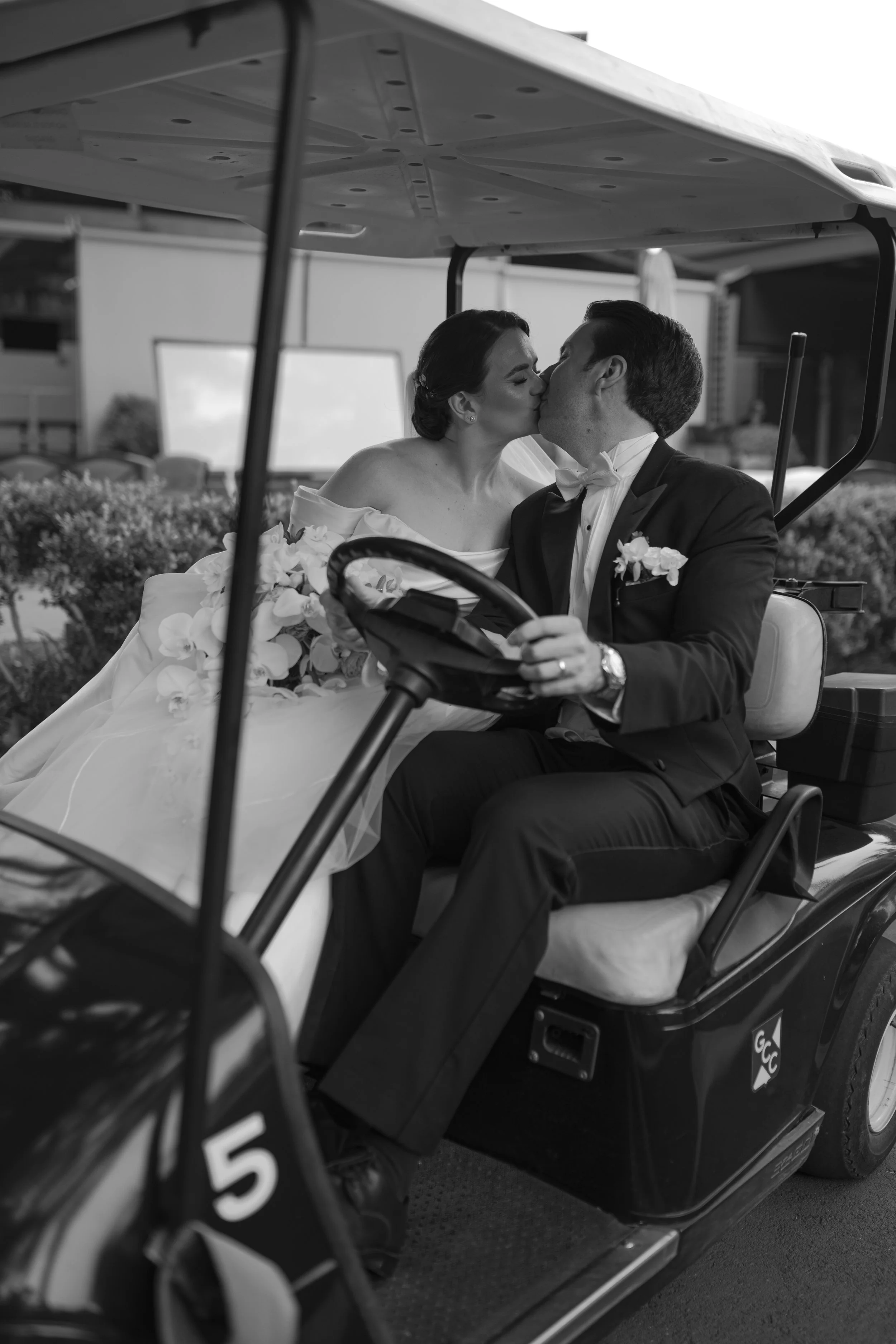 Una pareja de recién casados se besa en un carrito de golf, con la mujer usando un vestido de novia y el hombre con un traje formal, en un entorno exterior.