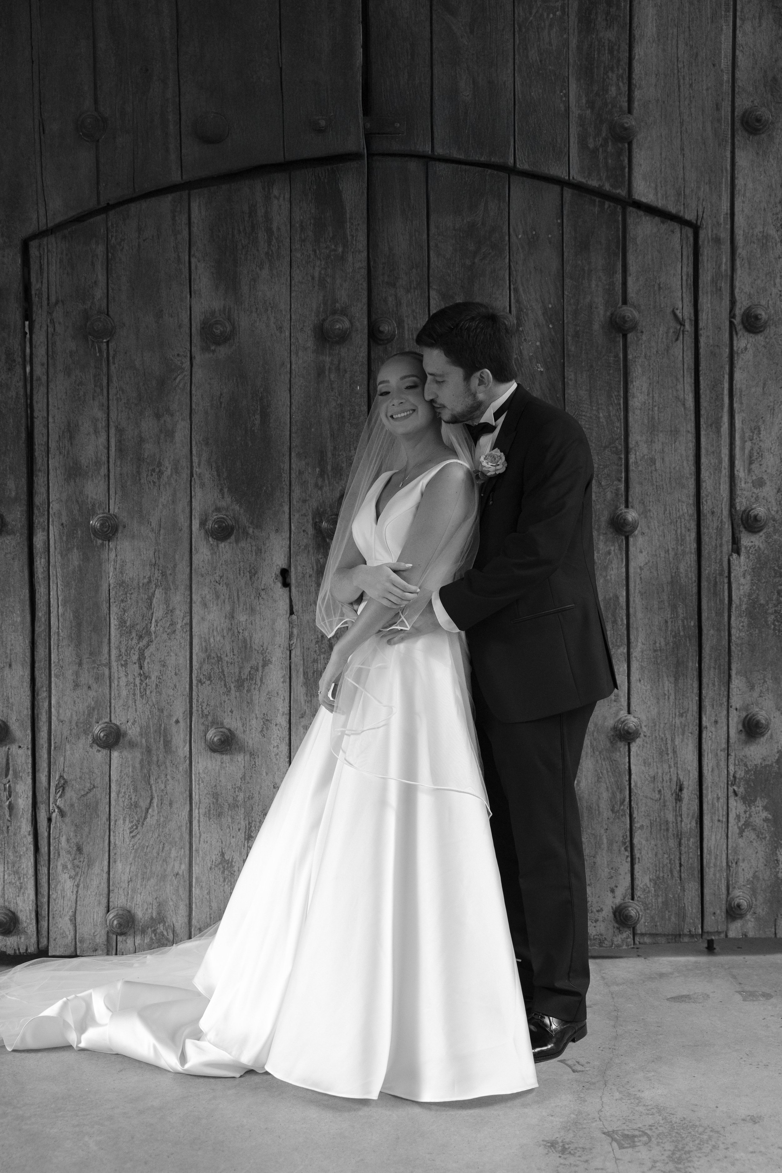 Una pareja de novios en su boda, con la mujer vestida con un vestido de novia y el hombre con un traje y corbata, posando juntos frente a una puerta de madera antigua.
