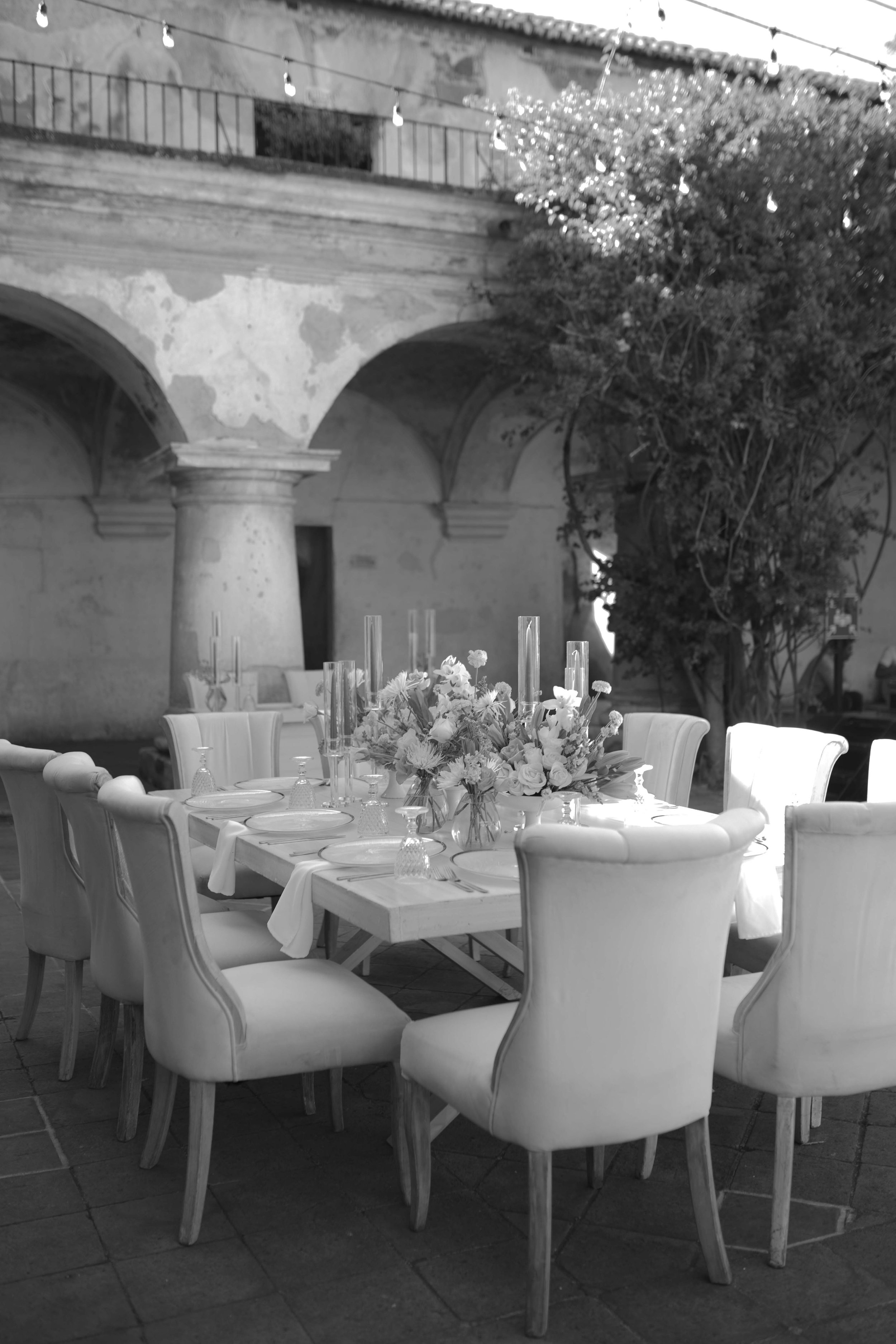 Mesa decorada con flores y velas en un patio con arquitectura colonial y paredes de piedra, en blanco y negro.