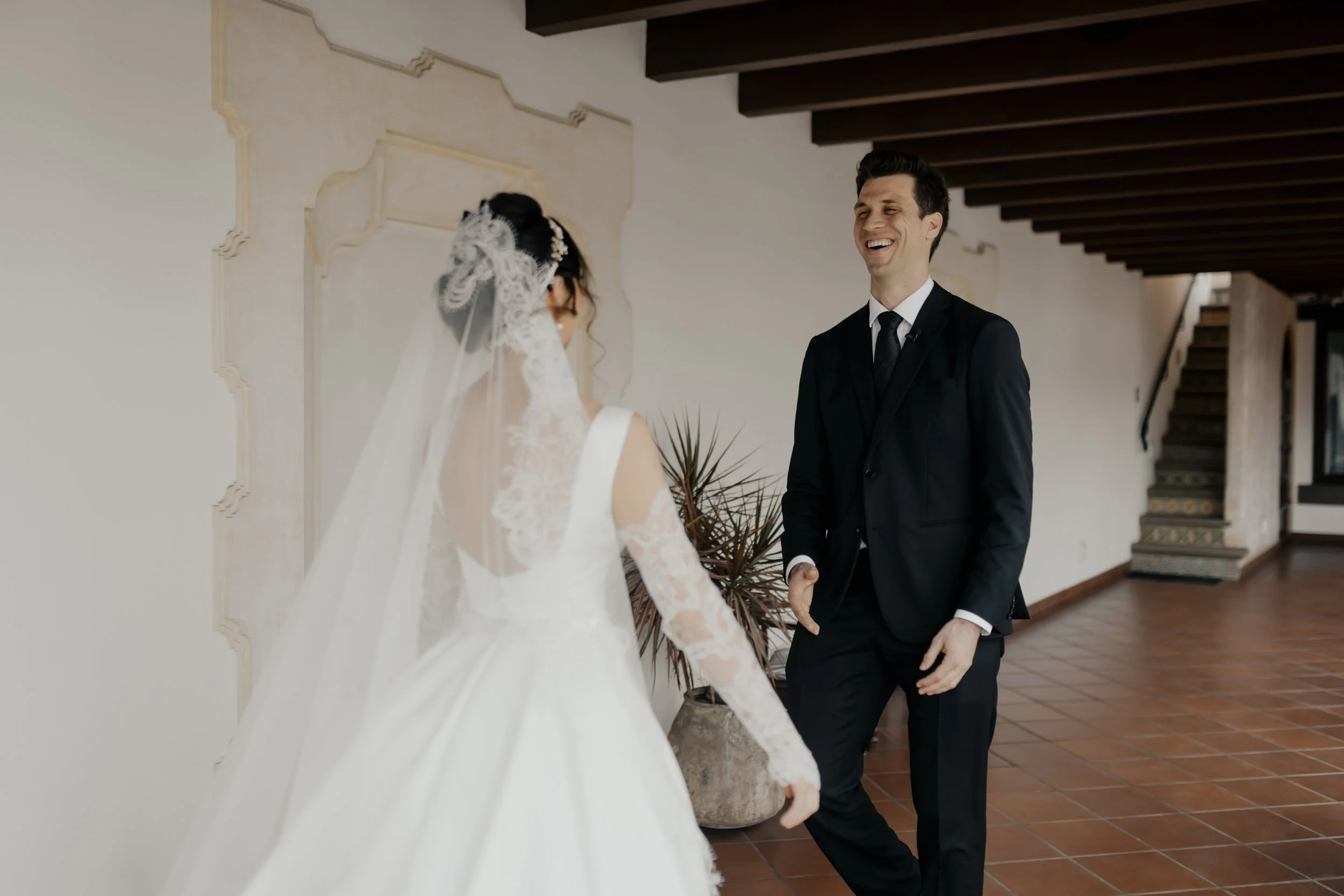 Pareja de recién casados riendo y caminando juntos en un ambiente interior elegante, ella con vestido de novia y velo, él con traje negro.