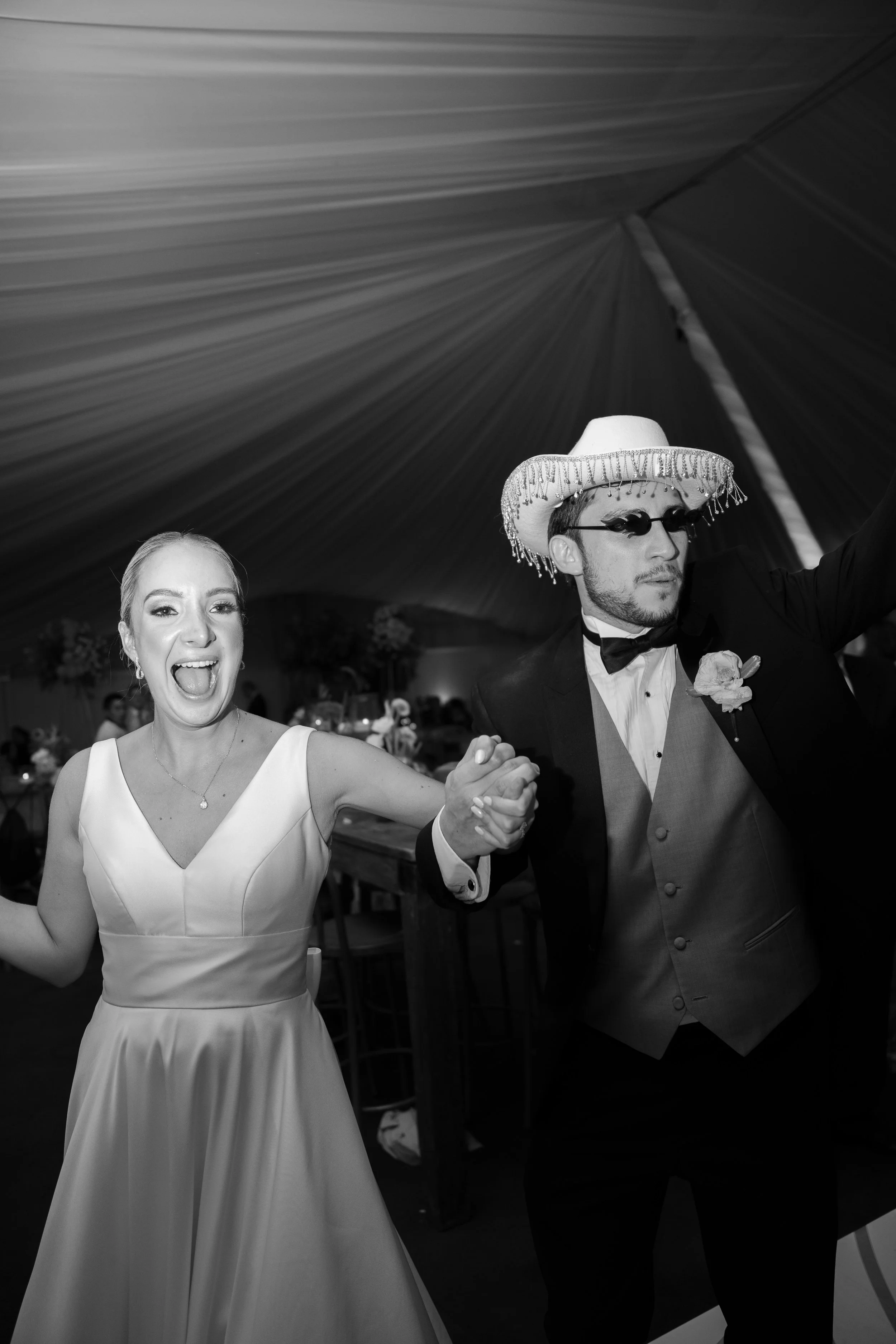 Pareja de novios bailando en una boda, la novia con vestido blanco y el novio con sombrero y gafas oscuras en un evento bajo una carpa.