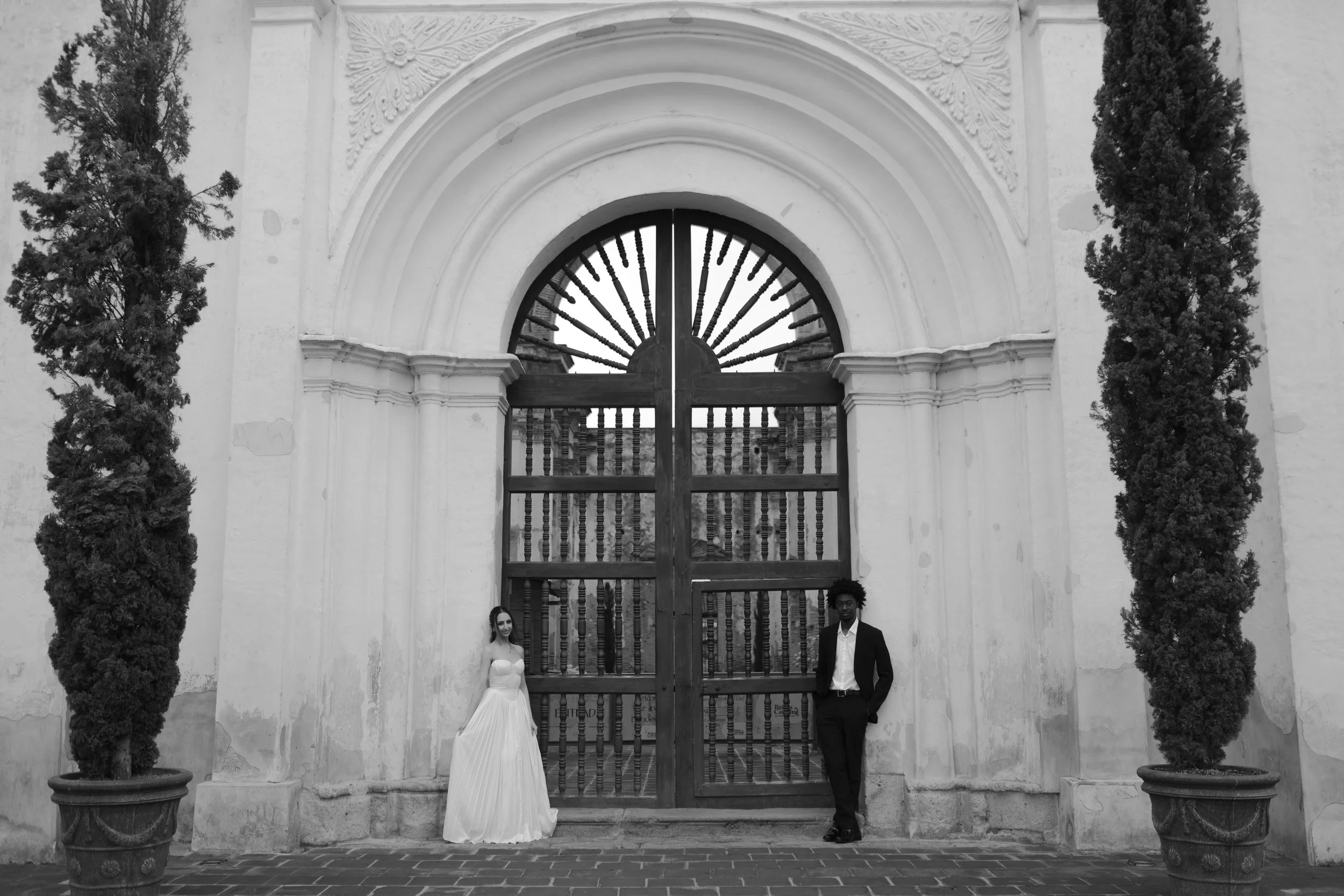Una pareja vestida con ropa formal, de pie a los lados de una gran puerta de madera decorada con barrotes, en la entrada de un edificio antiguo. La mujer lleva un vestido largo y la expresión de ambos es sonriente.