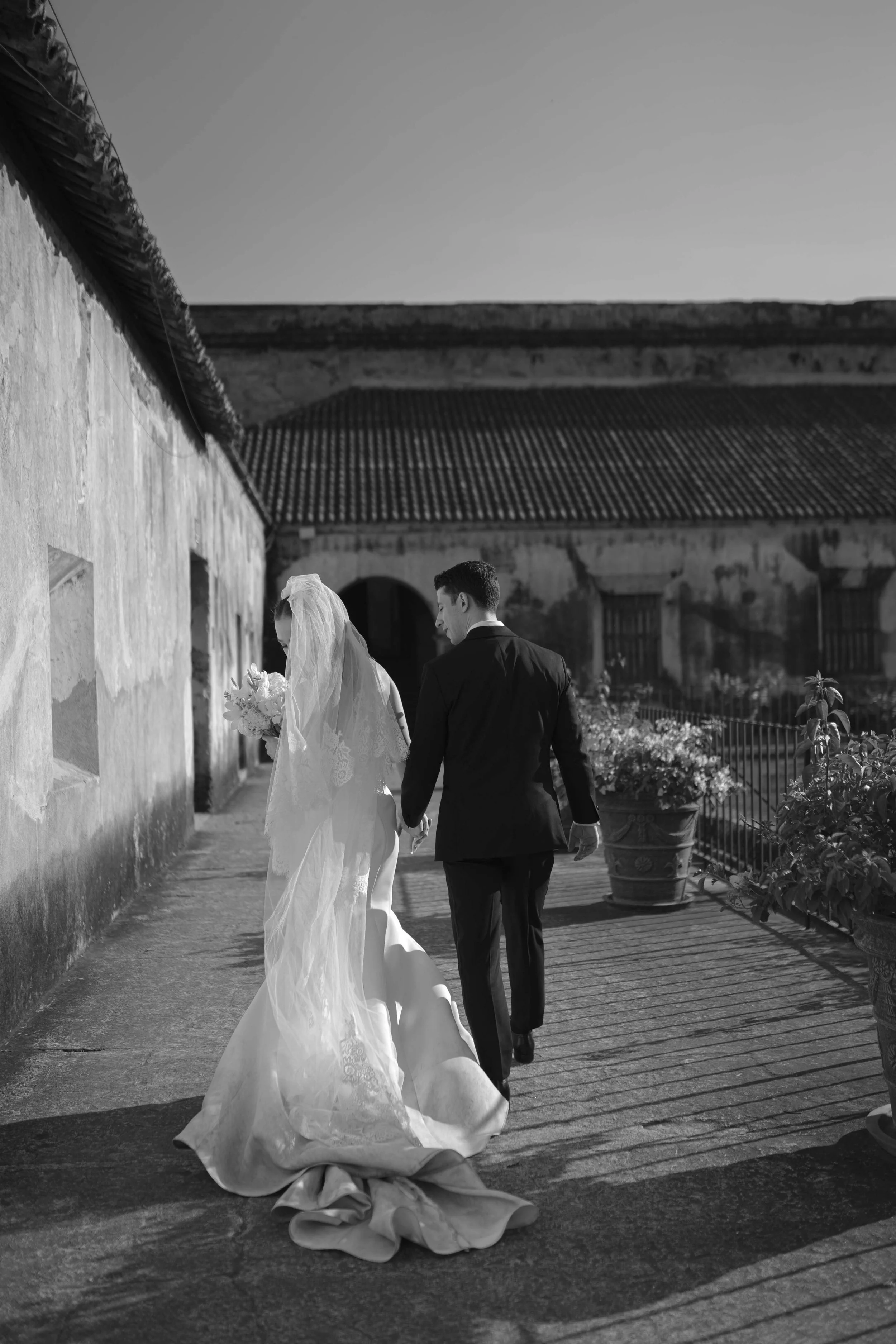Una pareja de recién casados caminando de la mano en un ambiente exterior, con la mujer vestida con un vestido de novia y un velo, y el hombre con traje oscuro, en un entorno con paredes antiguas y macetas con plantas.