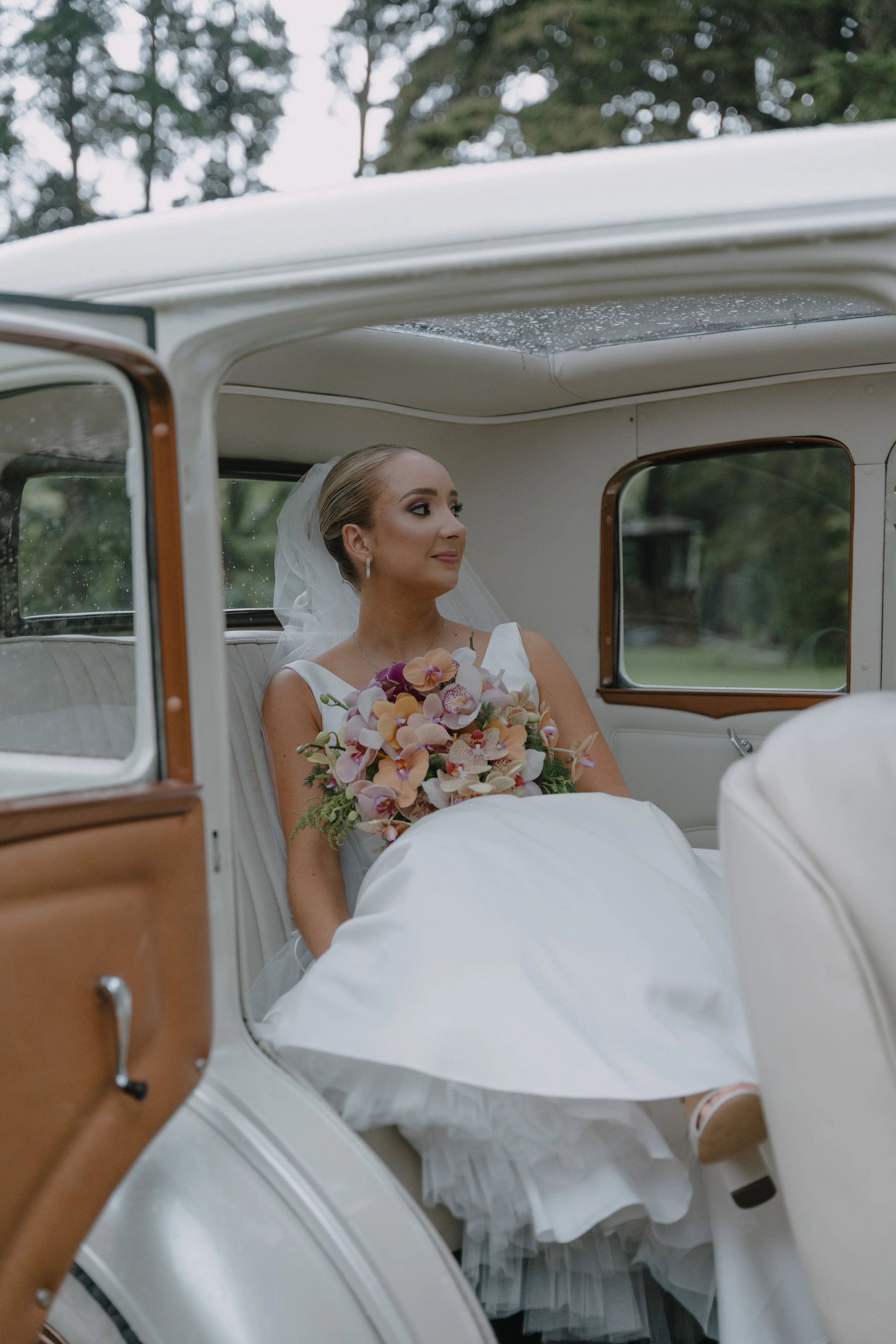 Una novia en vestido de boda y velo sentada en un coche clásico blanco, con ramo de flores, en un entorno exterior con árboles.