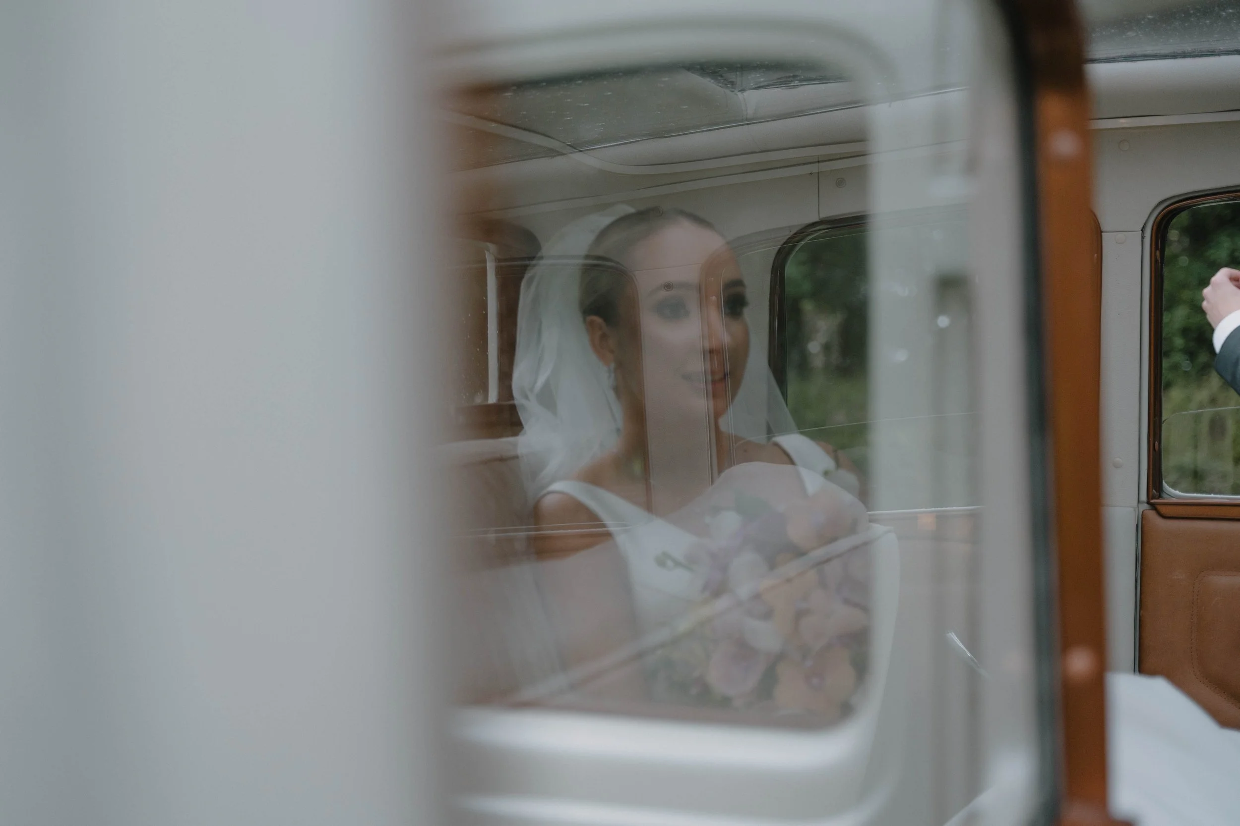 Mujer con vestido de novia y velo, reflejada en el cristal de una ventana de un autobús o vehículo clásico, que sostiene un ramo de flores.