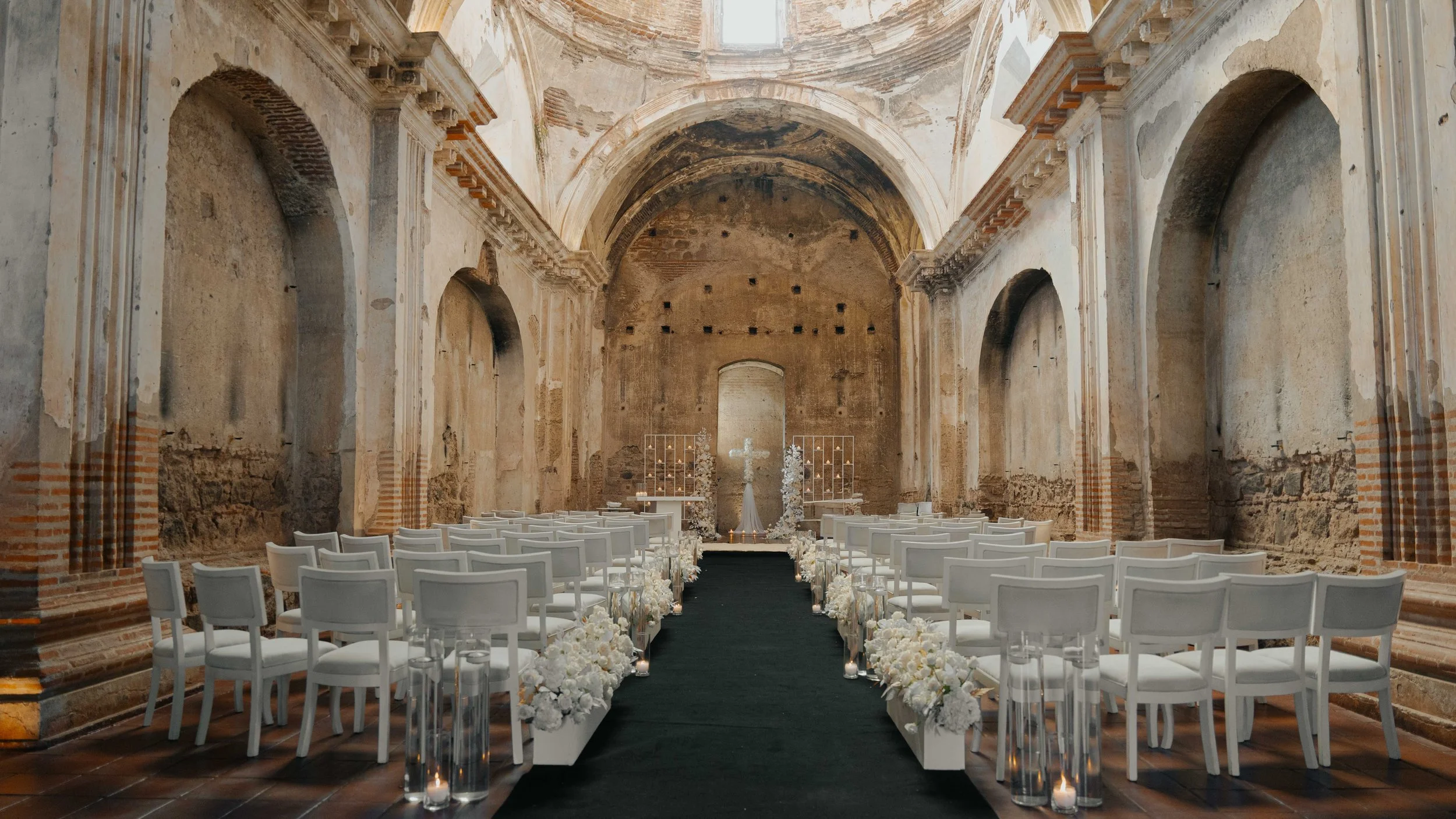 Interior de una iglesia antigua decorada para una boda, con sillas blancas en ambos lados de un pasillo cubierto con una alfombra negra, y un altar con una cruz y arreglos florales blancos.