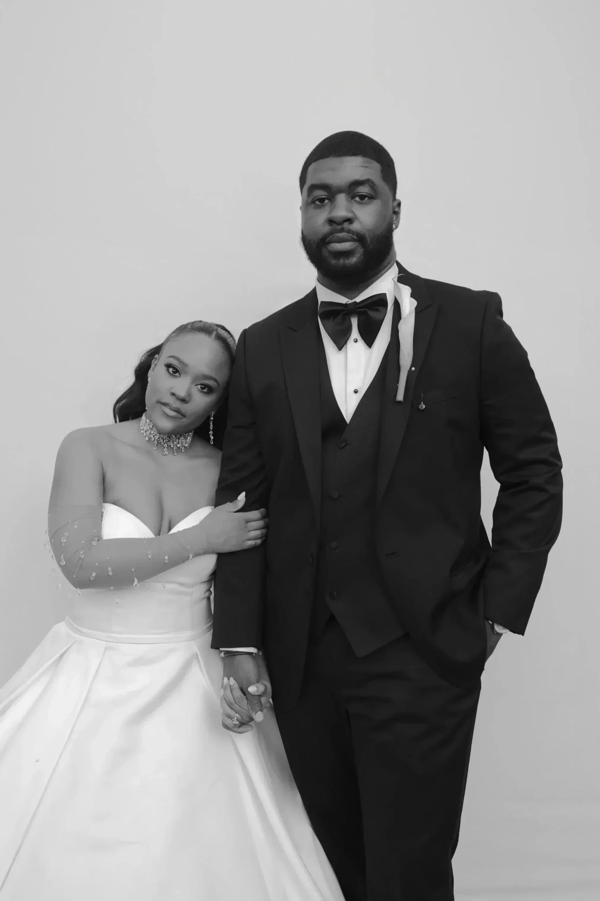 Una pareja vestida con ropa formal, la mujer con vestido de novia y el hombre con traje y pajarita, posando juntos en blanco y negro.