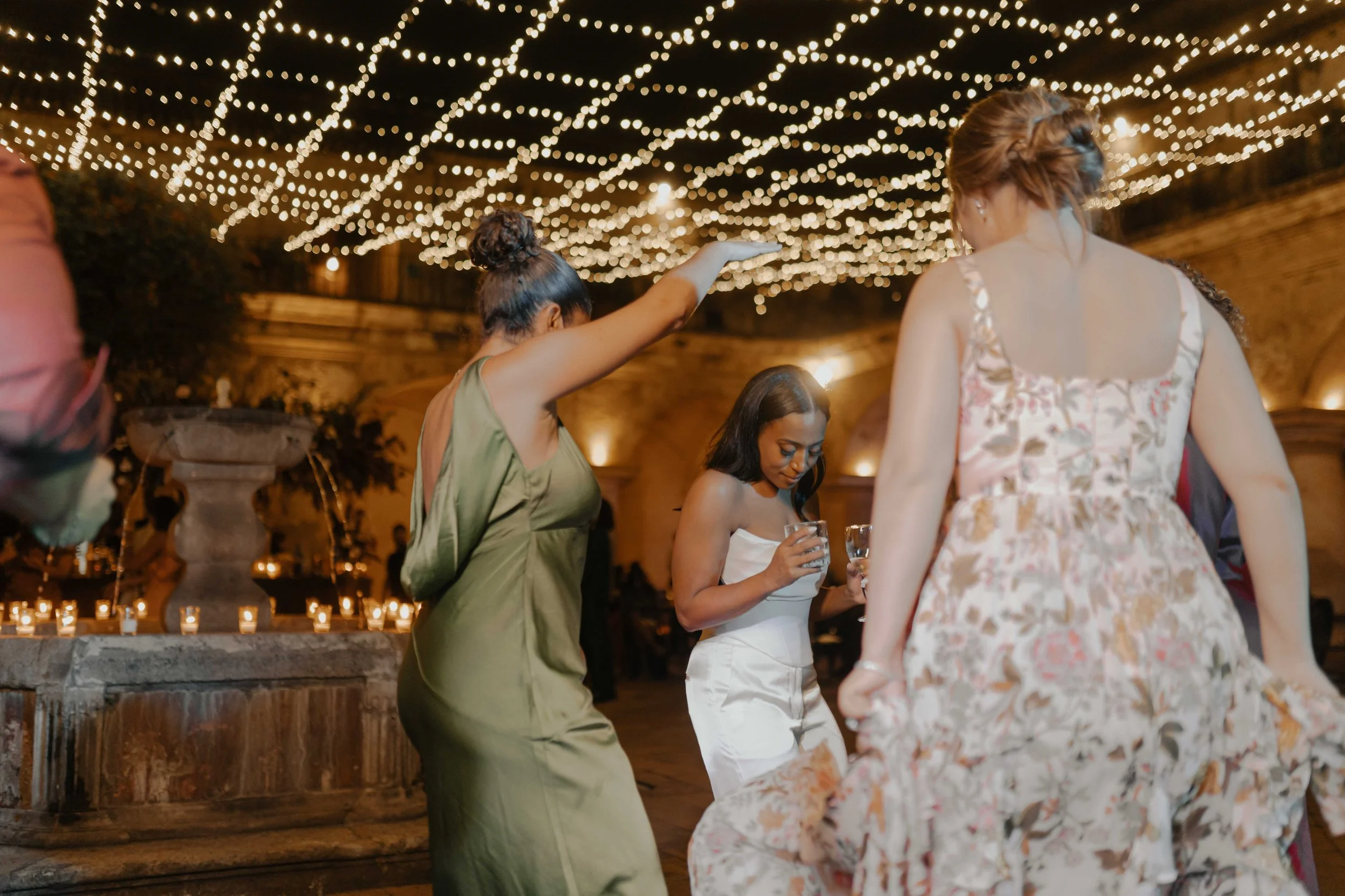 Personas bailando en una celebración por la noche, decorada con luces colgantes y velas en una fuente