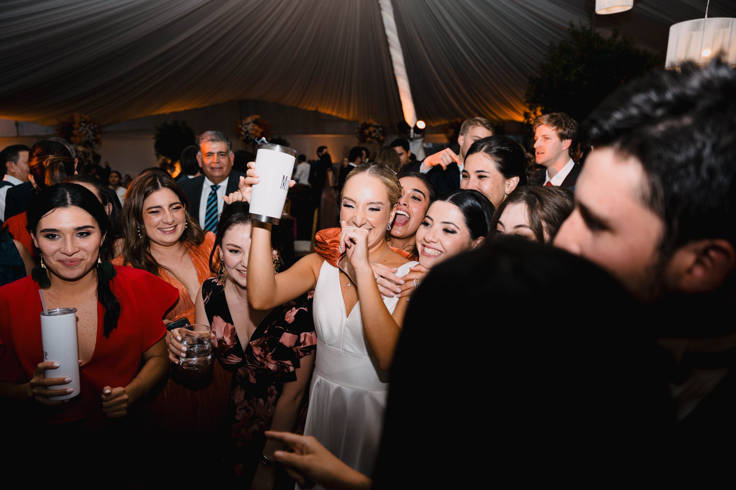 Grupo de personas sonriendo y divirtiéndose en una celebración en interiores, algunas sosteniendo bebidas en las manos.