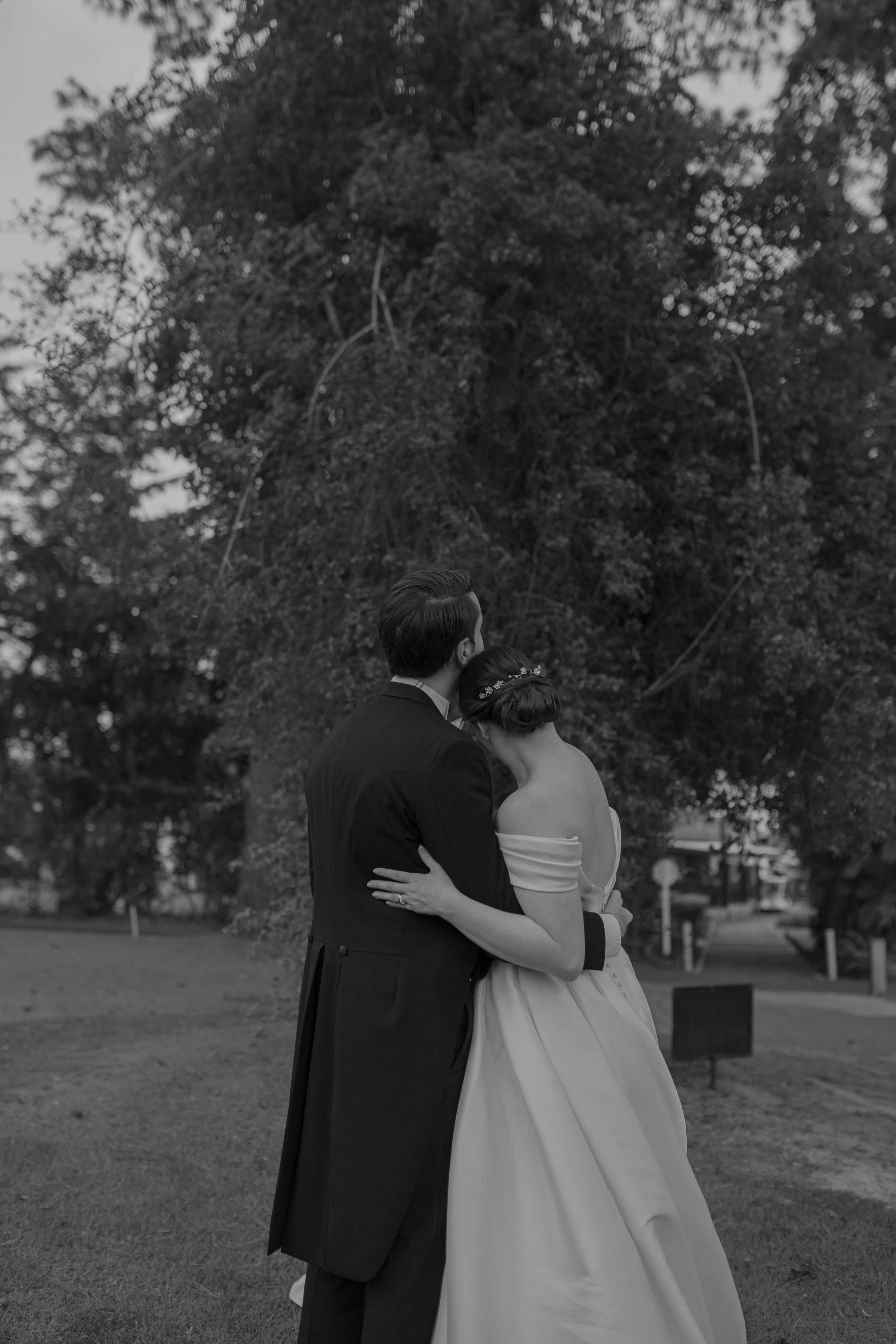 Pareja de recién casados abrazados, con el hombre vestiendo frac y la mujer con vestido de novia, en un parque con árboles grandes.