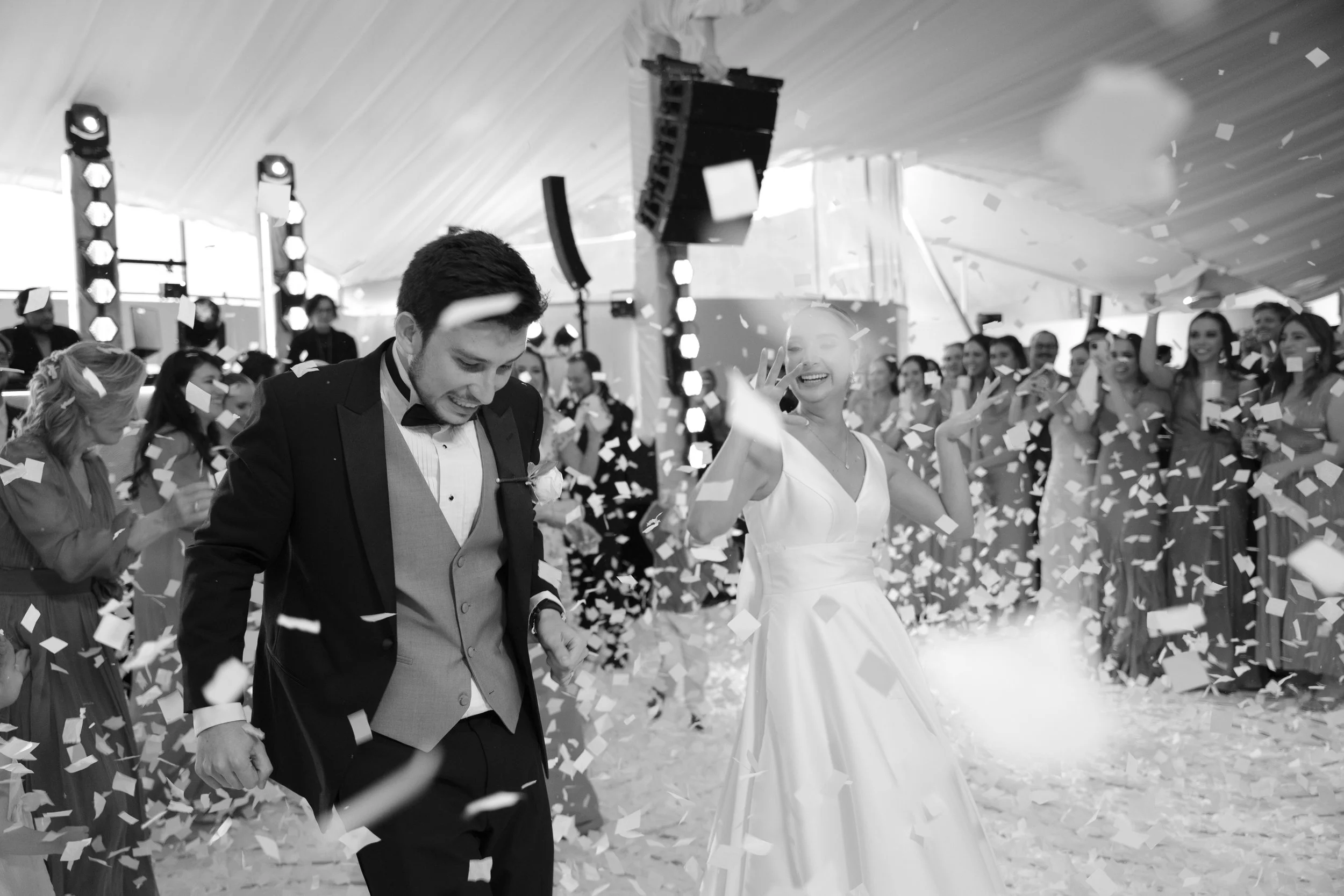 Una pareja de novios bailando en su boda rodeados de invitados mientras caen confeti y aplauden, en un ambiente festivo y alegre.