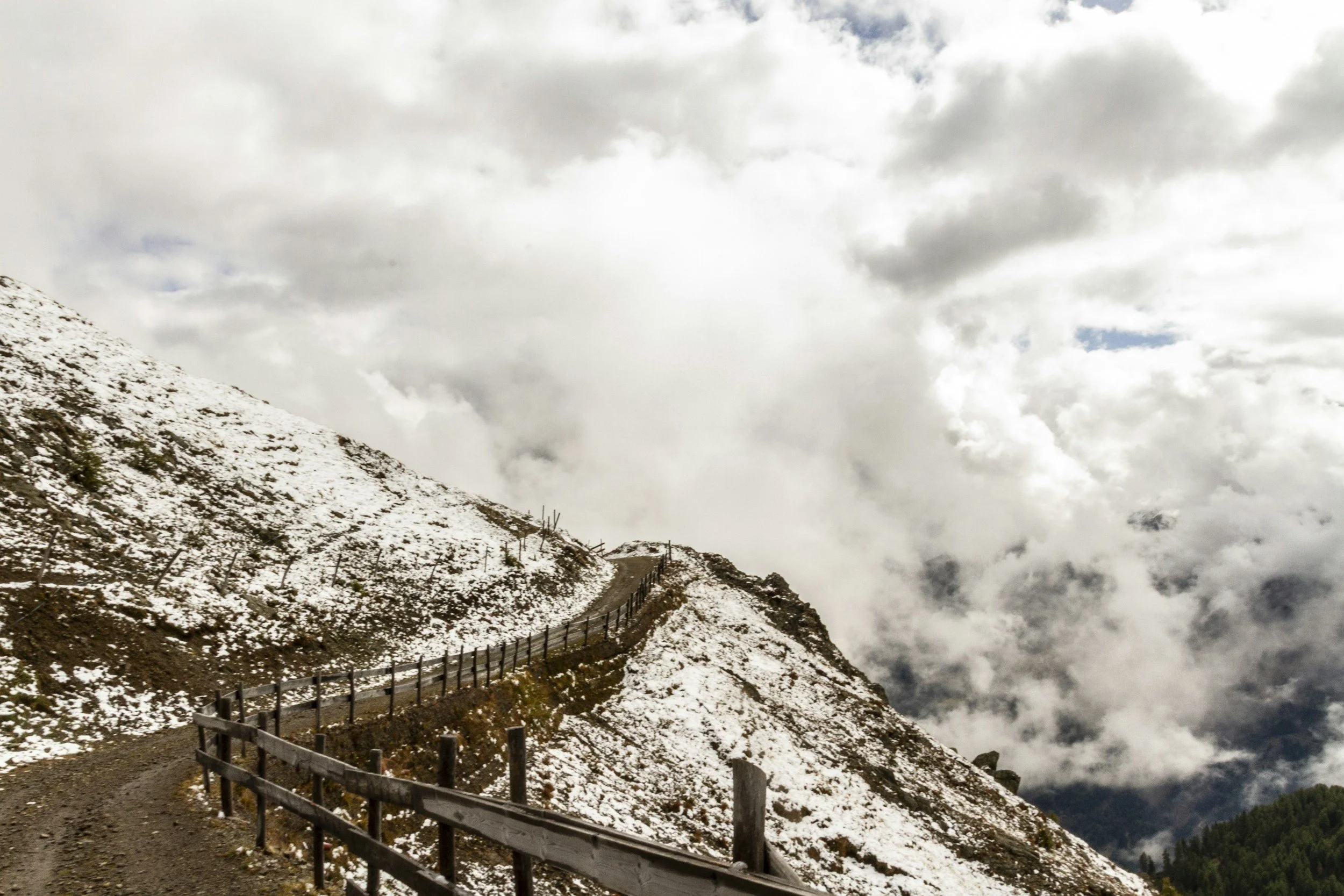 A narrow dirt path with a wooden fence winds along a snow-dusted mountain ridge, disappearing into thick clouds, suggesting a challenging yet purposeful journey forward.