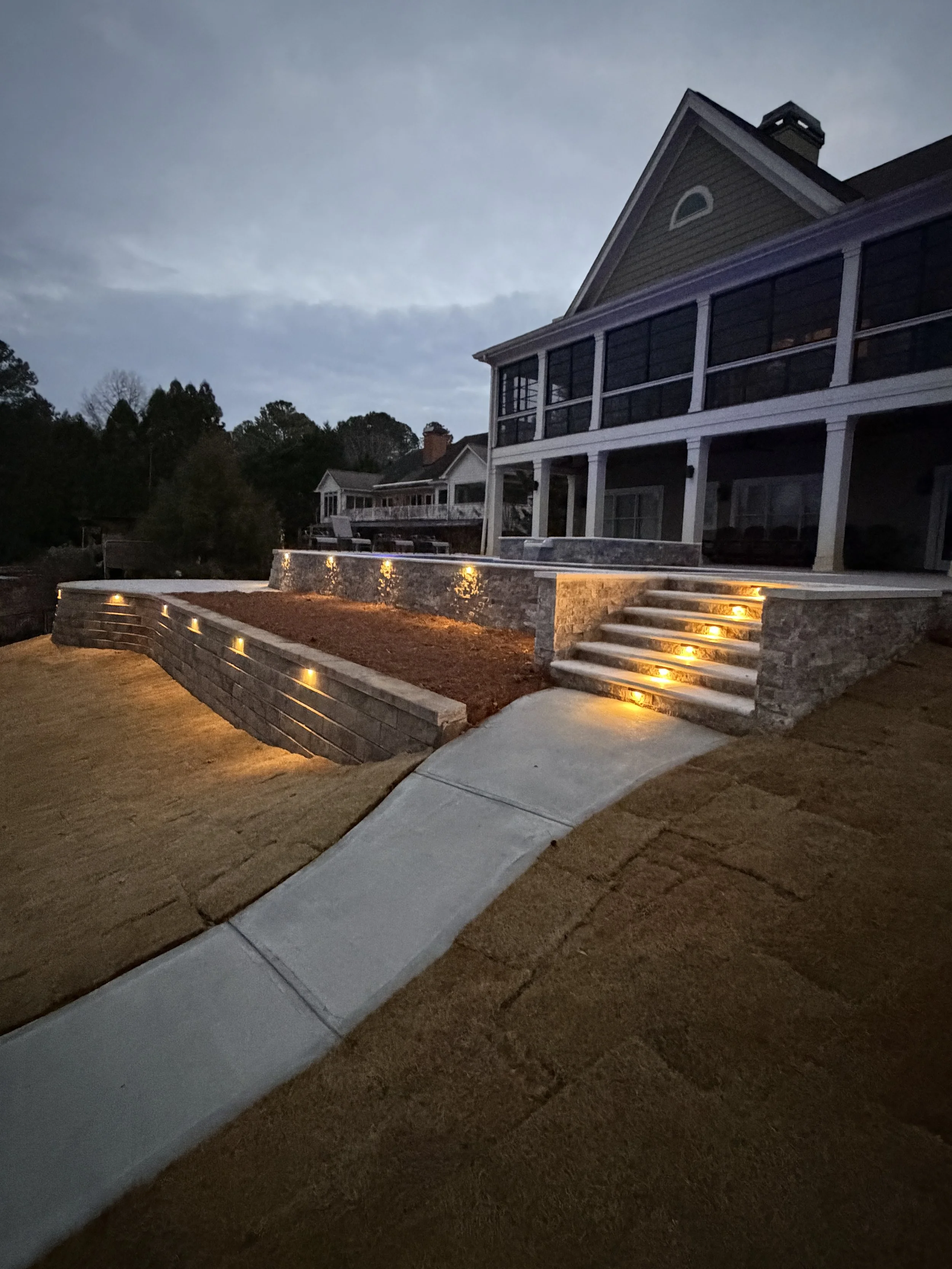 A residential house with a landscaped yard illuminated by outdoor lighting, including steps with lights and a curved pathway leading to the house.