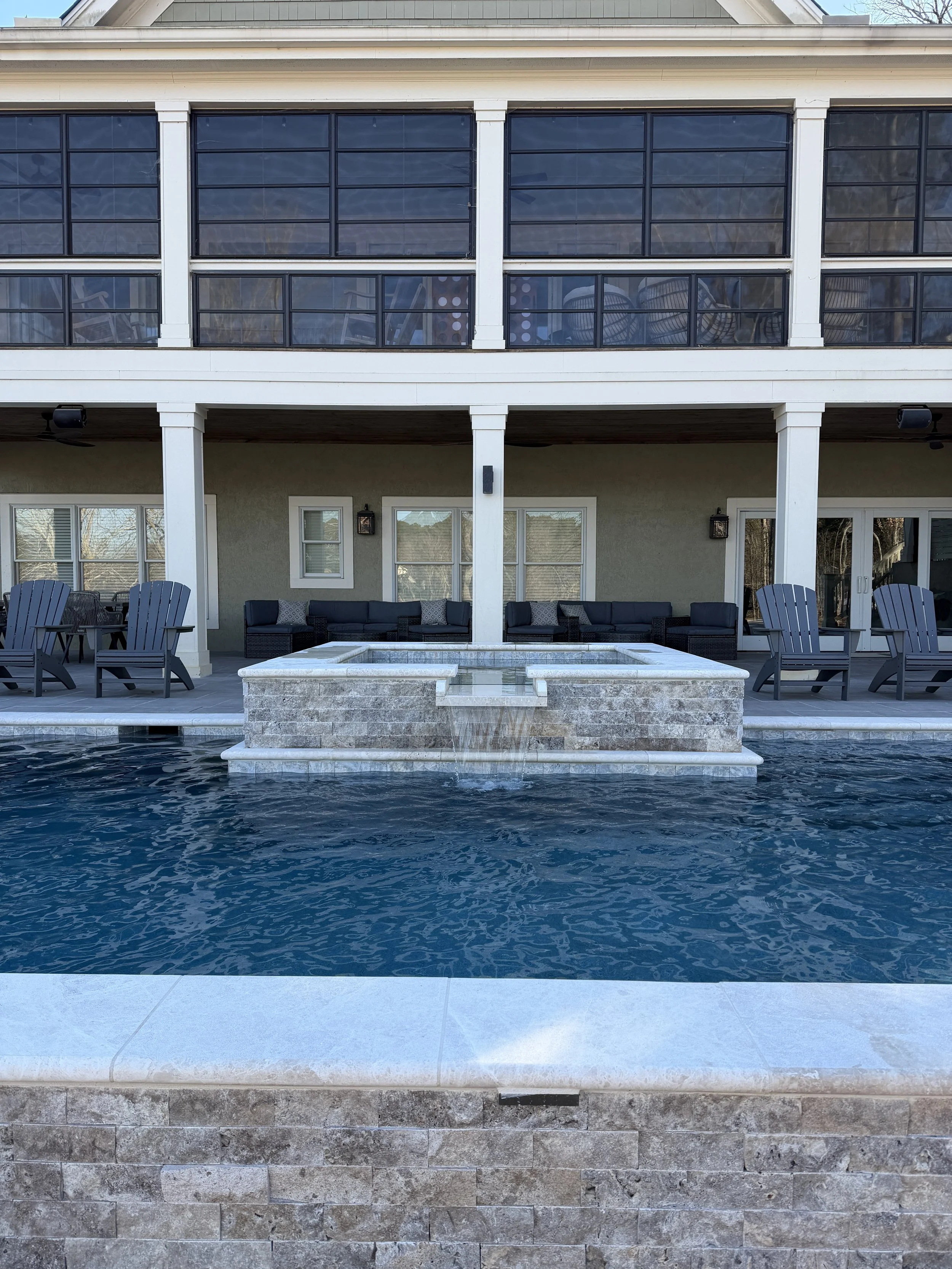 Outdoor pool with a small waterfall feature, surrounded by lounge chairs, with a house or building in the background that has a balcony and large windows.
