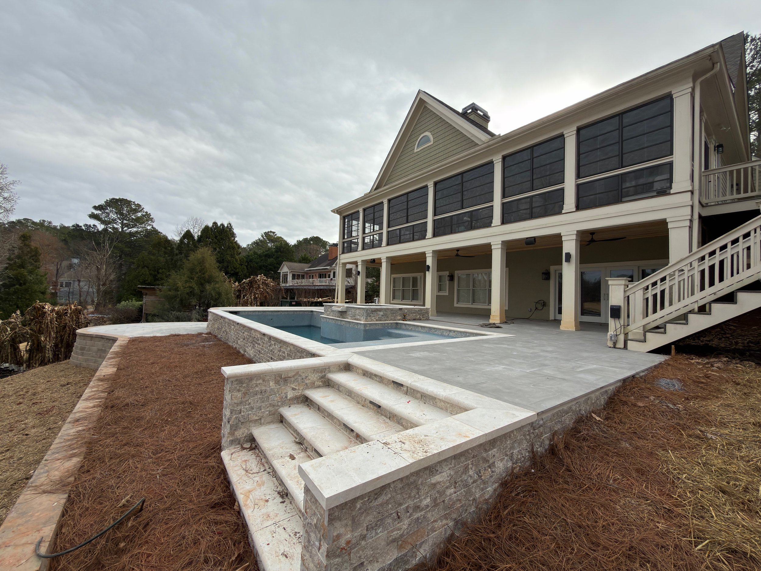 Backyard view of a two-story house with a patio, a modern pool, and a staircase leading up to the deck, during cloudy weather.