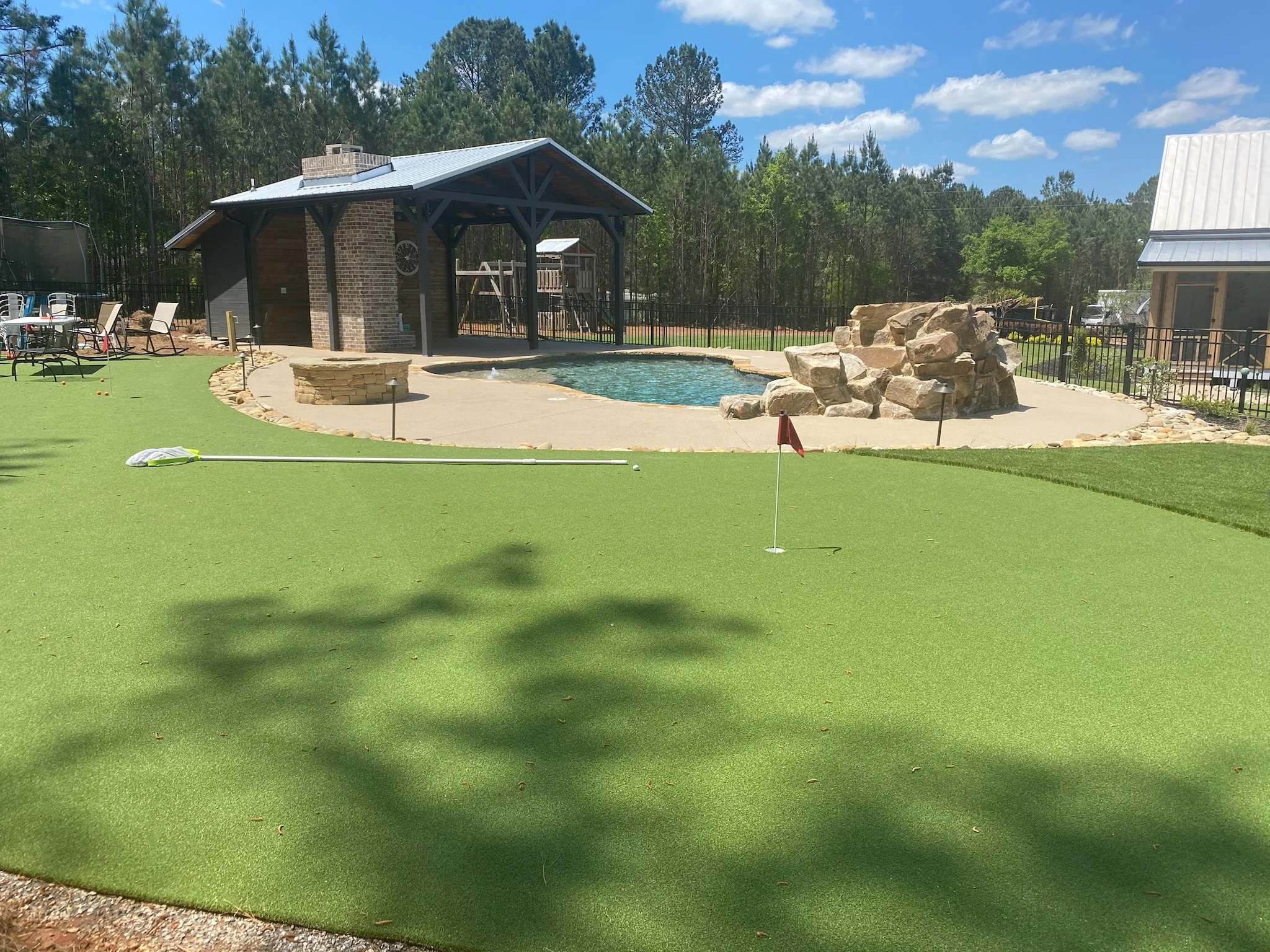 A backyard with a putting green, a pool with a rock waterfall, a covered pavilion, and outdoor seating on a sunny day.
