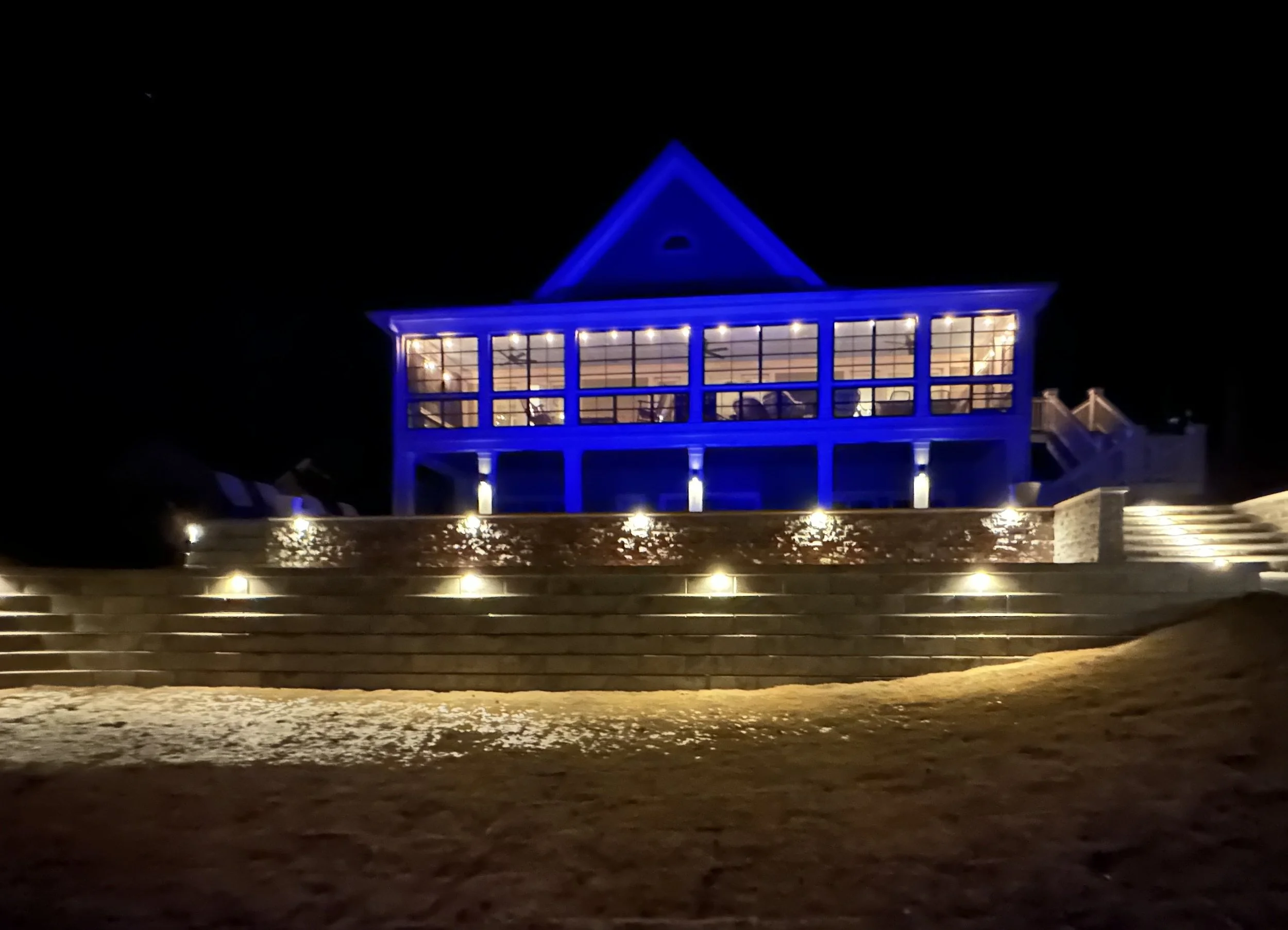 A house illuminated with blue and warm white lights at night, with a staircase and a water feature in front.