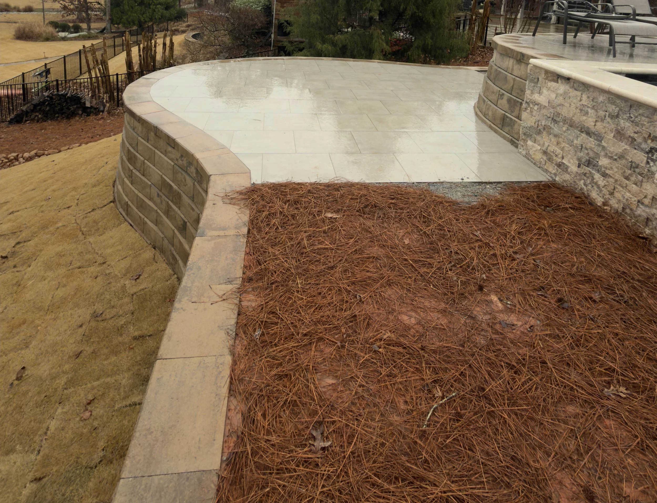A newly paved outdoor patio with a curved stone wall, a section of reddish-brown pine needles on damp ground, and a yellowish lawn with patches of dirt.
