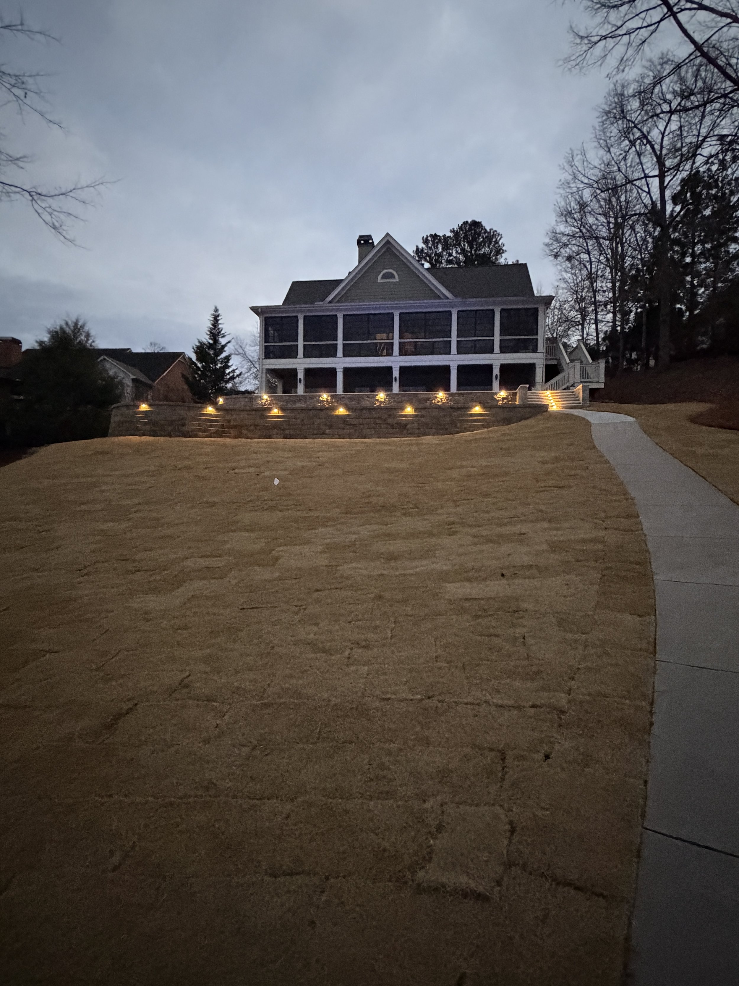 A large house at dusk with a screened porch and multiple levels, situated on a sloped lawn with steps and a curved walkway, and exterior lighting along the retaining wall.