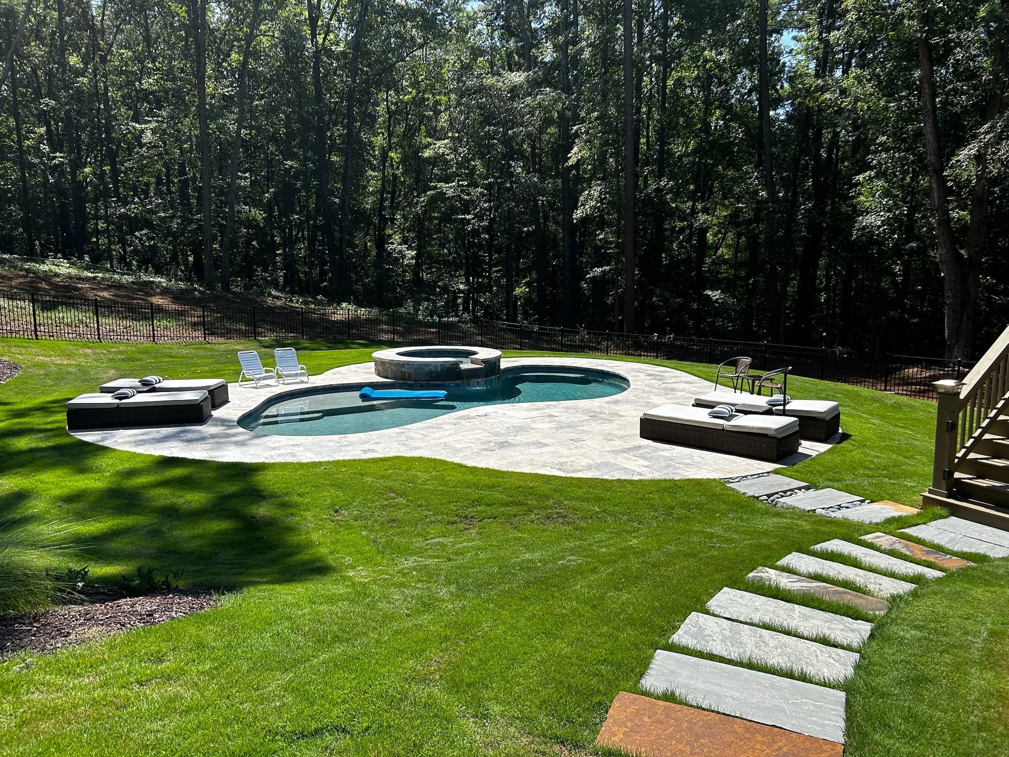 Backyard with a pool, hot tub, lounge chairs, outdoor furniture, and a grassy area with stone stepping stones, surrounded by trees.