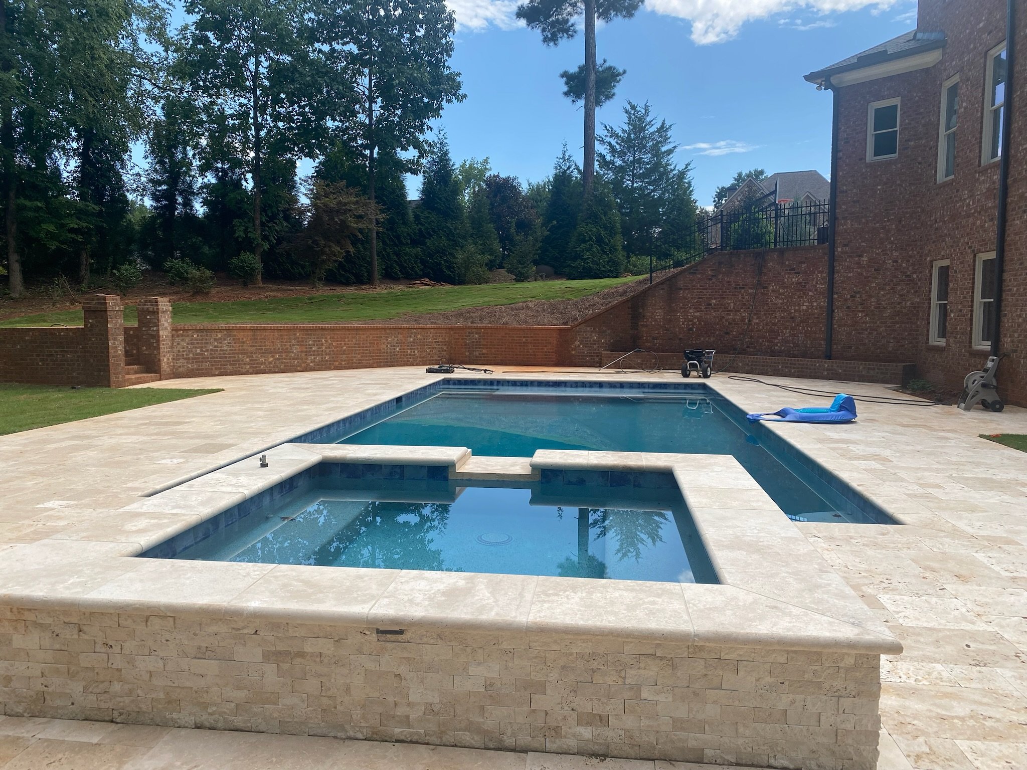 Backyard with a swimming pool and hot tub, surrounded by a beige stone patio, brick wall, trees, and a house in the background.