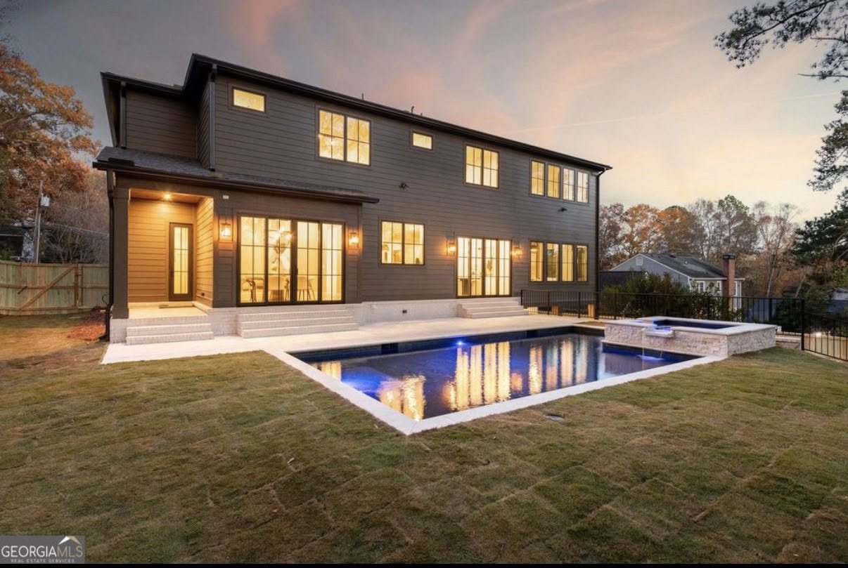 A modern two-story house with a backyard pool, surrounded by a grassy lawn and a wooden fence, during sunset with lit windows illuminating the home.