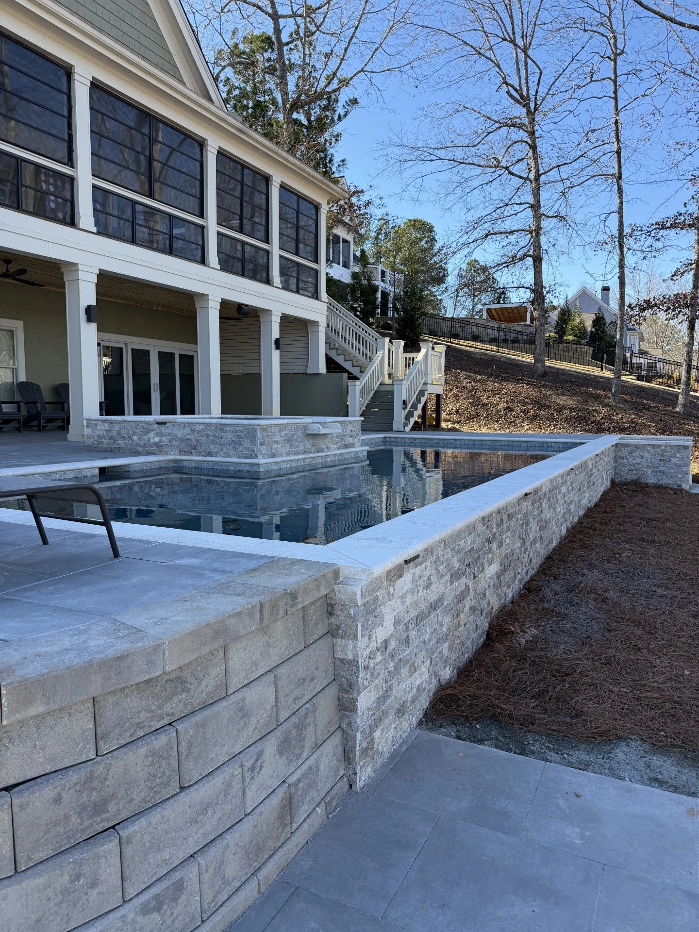 Modern backyard with a swimming pool, stone retaining wall, and a large multi-story house with screened porch and staircase.