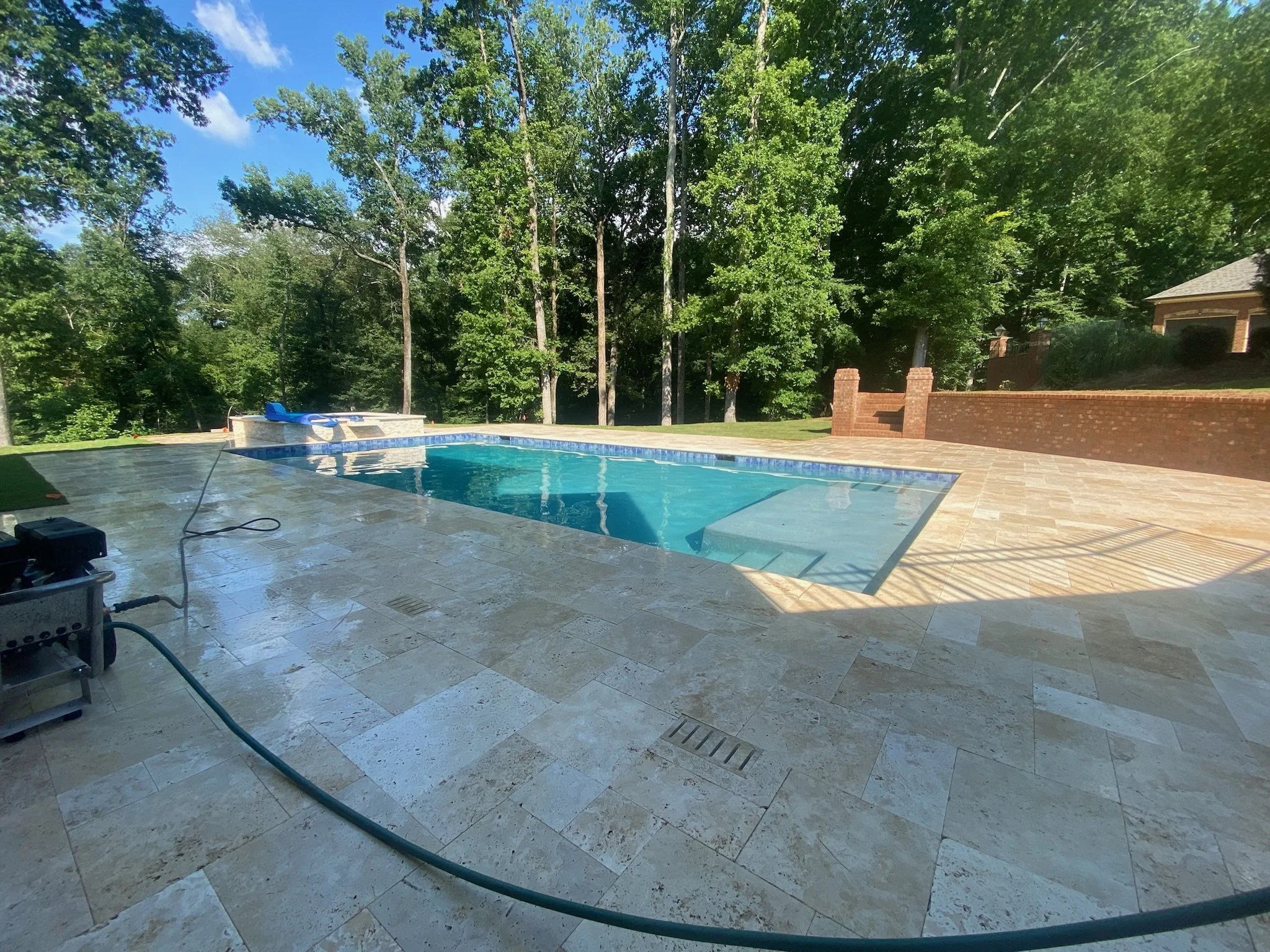 Backyard swimming pool with beige tile surround, surrounded by green trees and a brick wall, on a sunny day.