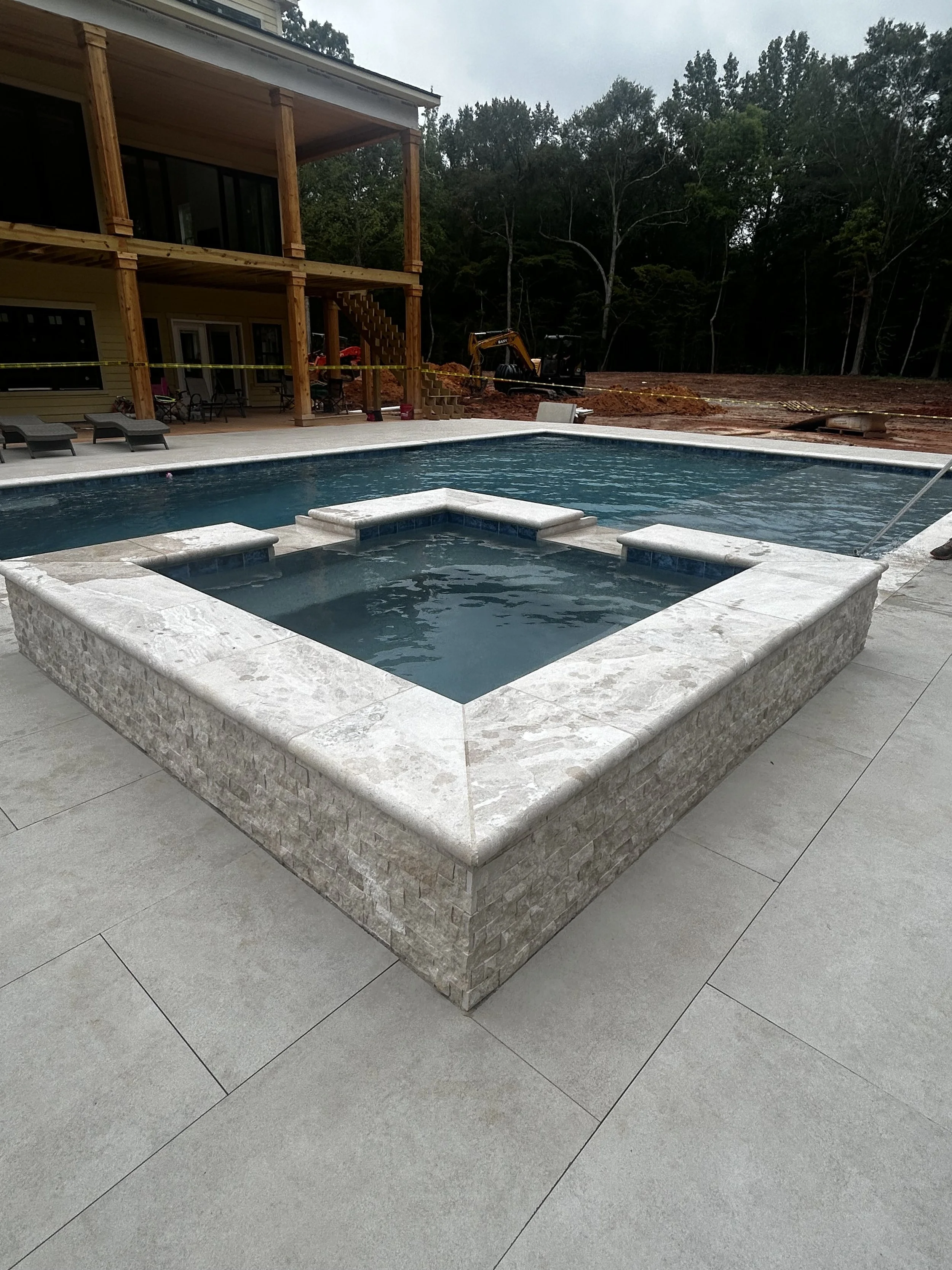 Newly constructed backyard pool with a hot tub, surrounded by a stone border and concrete patio, with a partially built house and construction equipment in the background.