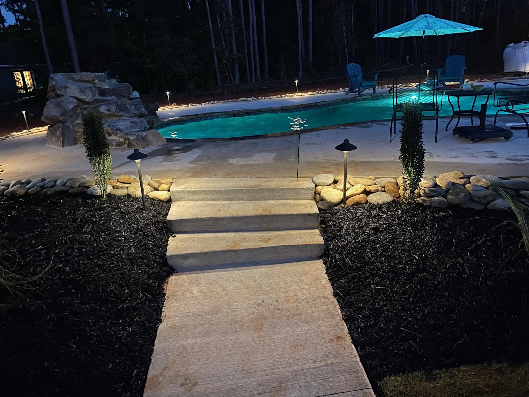Nighttime view of a backyard with a lit swimming pool, stone waterfall feature, outdoor furniture with an umbrella, and decorative lighting along the pathway.